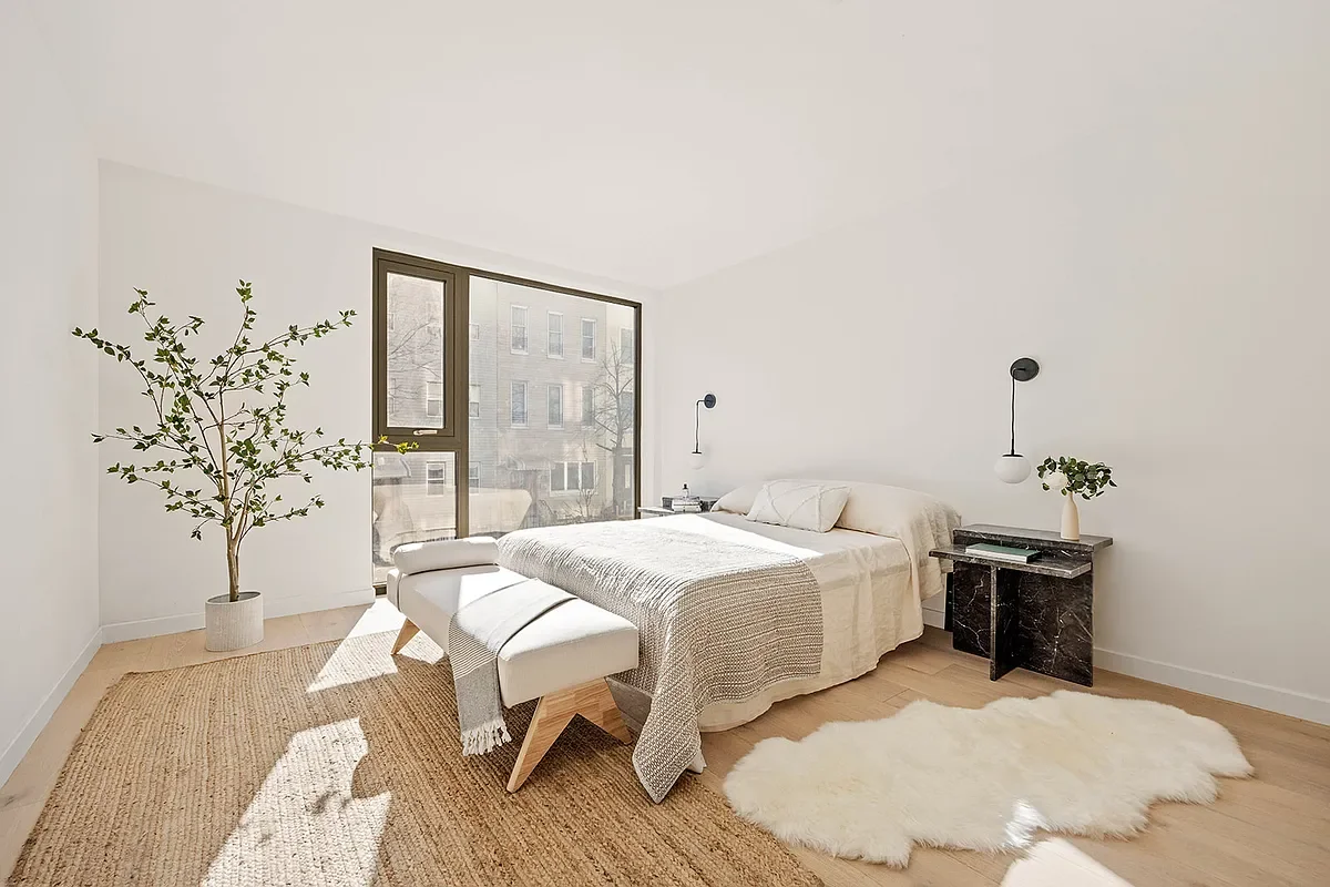 Bright, minimalist bedroom with a large window, potted plant, neutral-colored bedding, black marble nightstand, and white plush rug. Developed by Ten Talents Management.