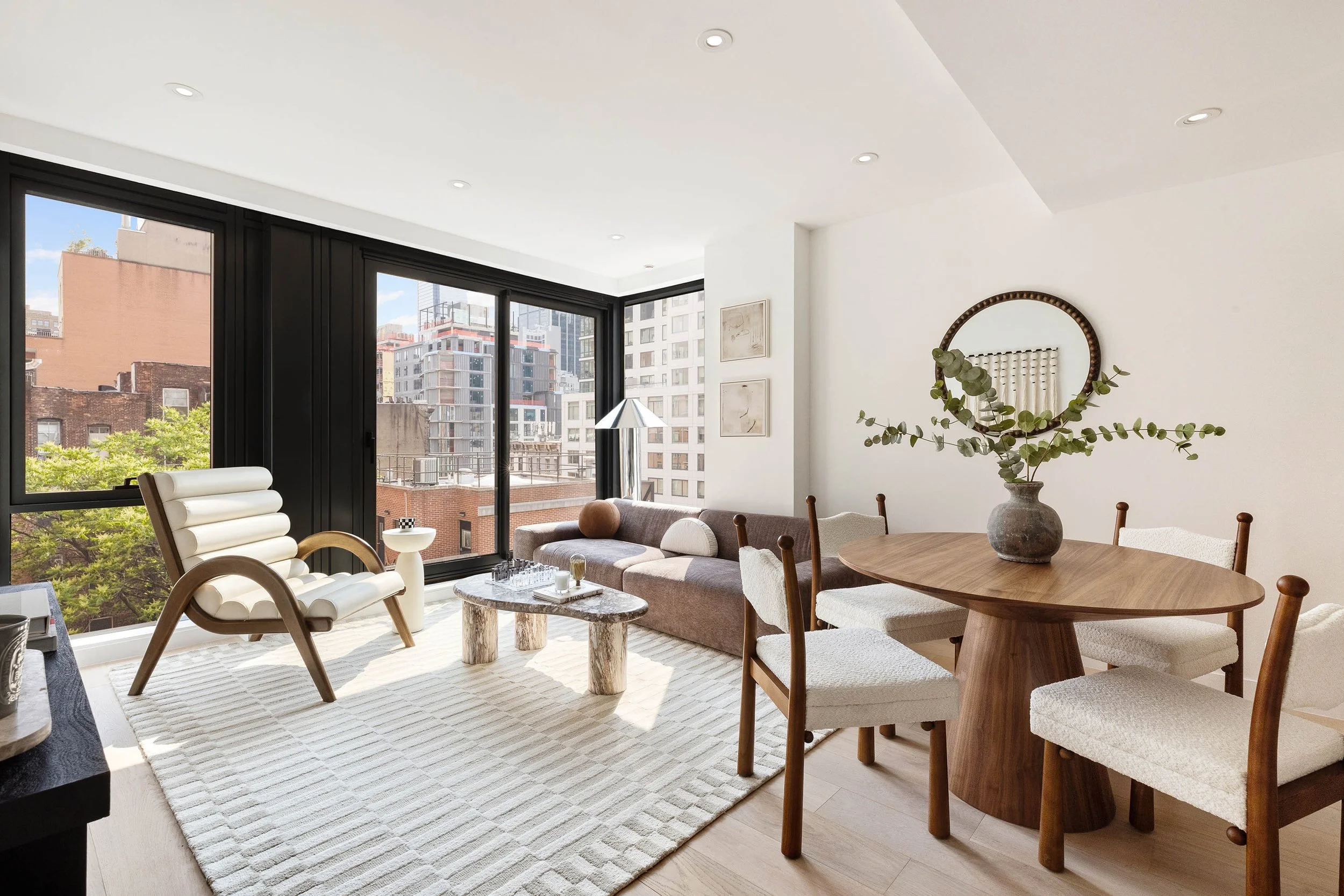 Modern living room with large windows showing city buildings, furnished with a white lounge chair, a beige sofa, a wooden dining table with white chairs, a round mirror with greenery, and a marble coffee table. Developed by Ten Talents Management.