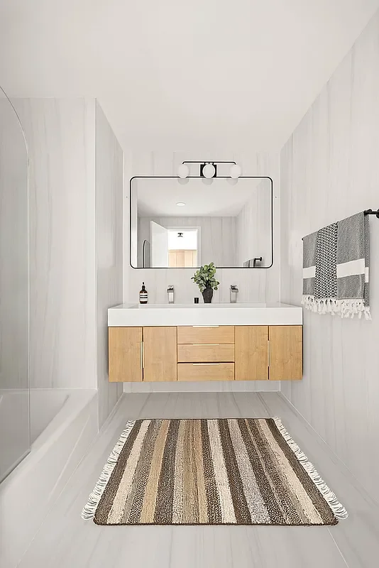 Modern bathroom with a floating wood vanity, large mirror, potted plant, and striped rug on light-colored flooring. Developed by Ten Talents Management.