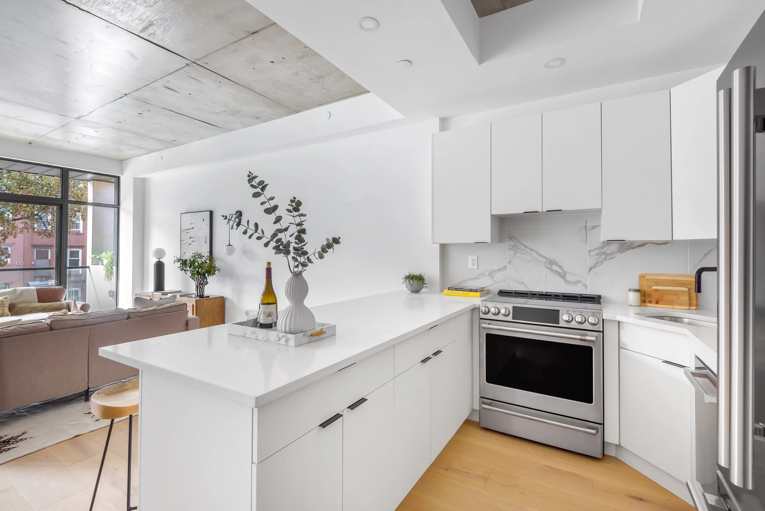 Modern kitchen with white cabinetry, marble backsplash, stainless steel oven, and a white countertop with decorative vases and plants. Developed by Ten Talents Management.