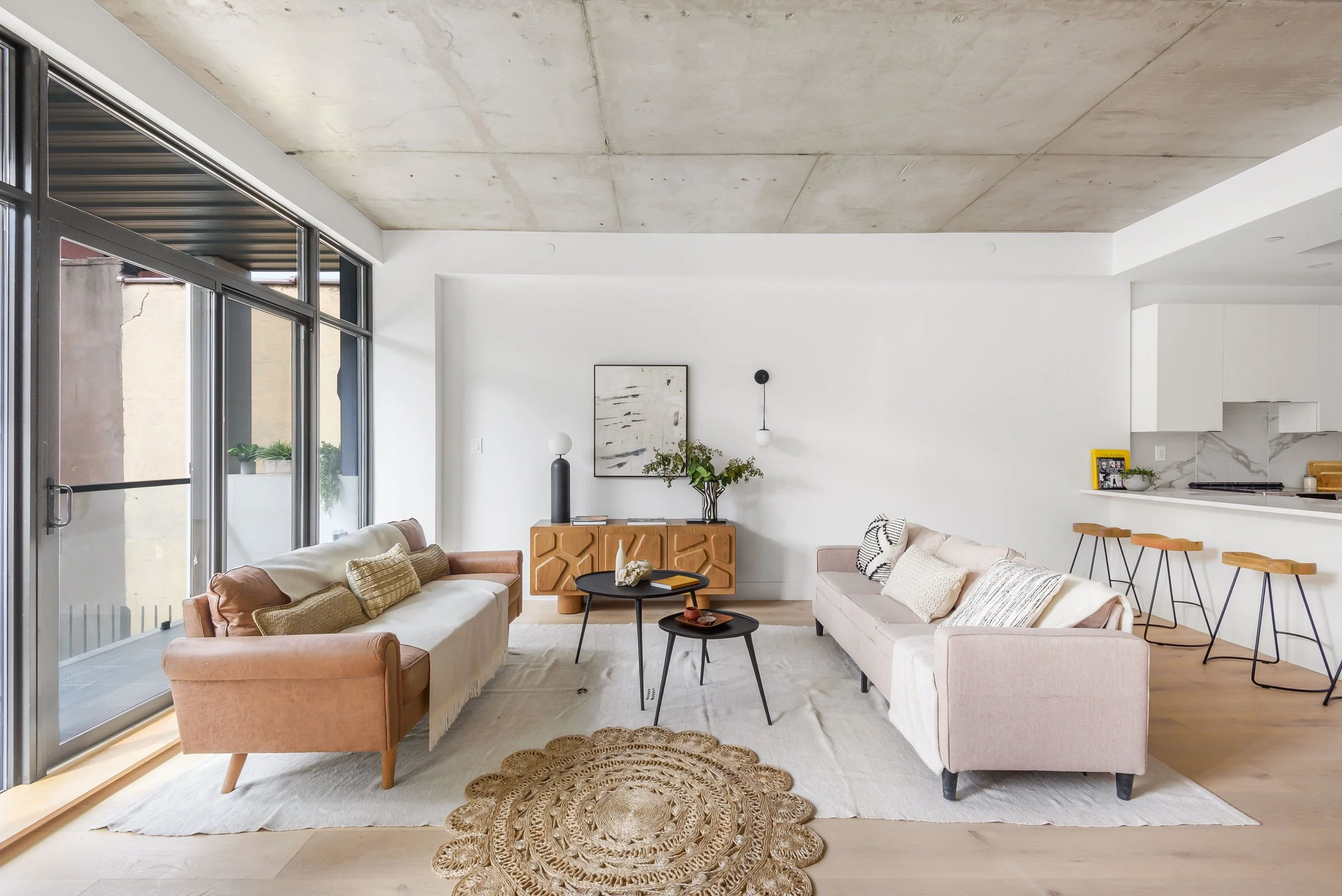 Living room with large glass sliding doors, pink and beige sofas, a black coffee table, artwork on the wall, and a kitchen area in the background. Developed by Ten Talents Management.