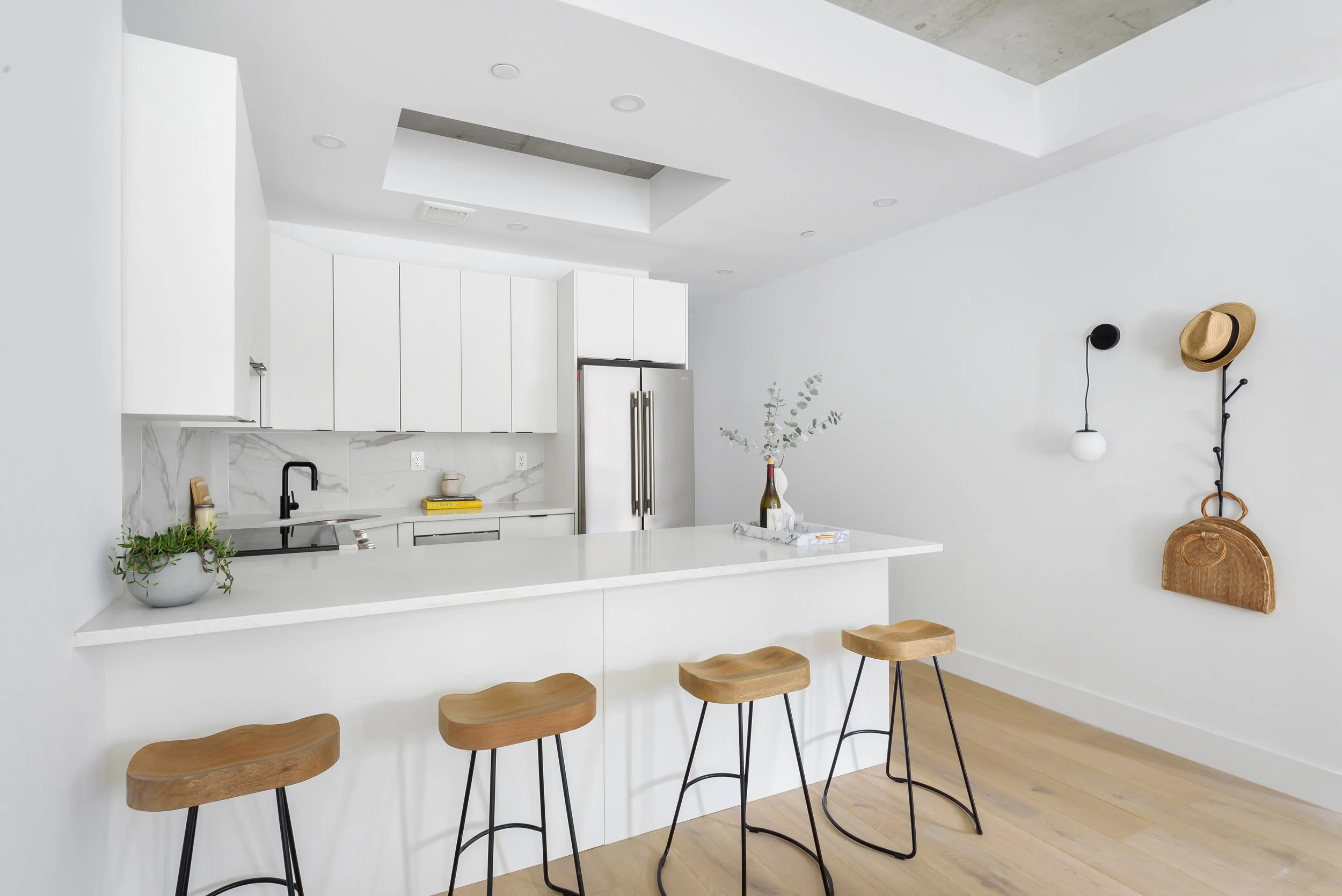 Modern kitchen with white cabinets, marble backsplash, and bar seating under concrete roof. Decor includes a potted plant, a book, a vase with eucalyptus, and a hat on hooks. Developed by Ten Talents Management.