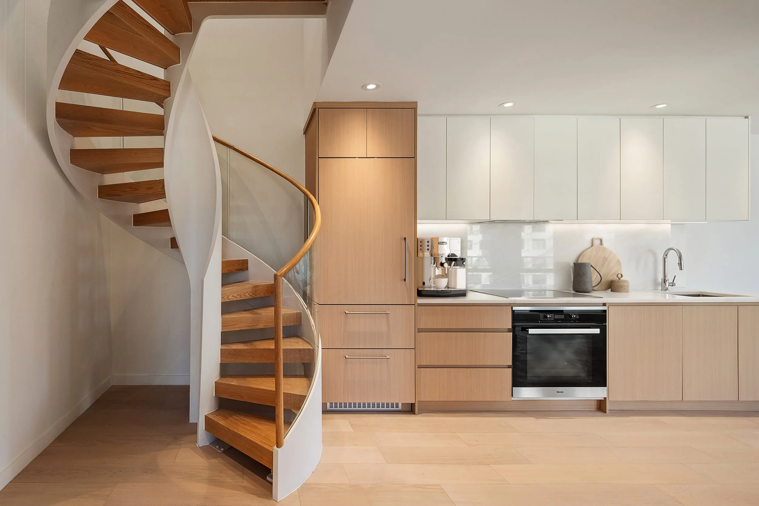 Interior view of a modern kitchen with a spiral wooden staircase to the left, light wood cabinets, white backsplash, and a countertop with a coffee machine, pitcher, and bowl. This is a kitchen developed by Ten Talents Management.
