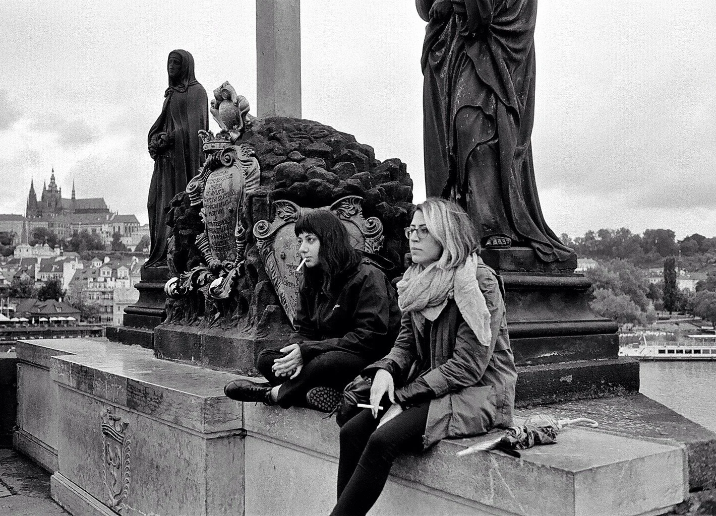 Two women sit on a ledge near ornate statues on a bridge, smoking cigarettes, with a cityscape and a castle in the background.