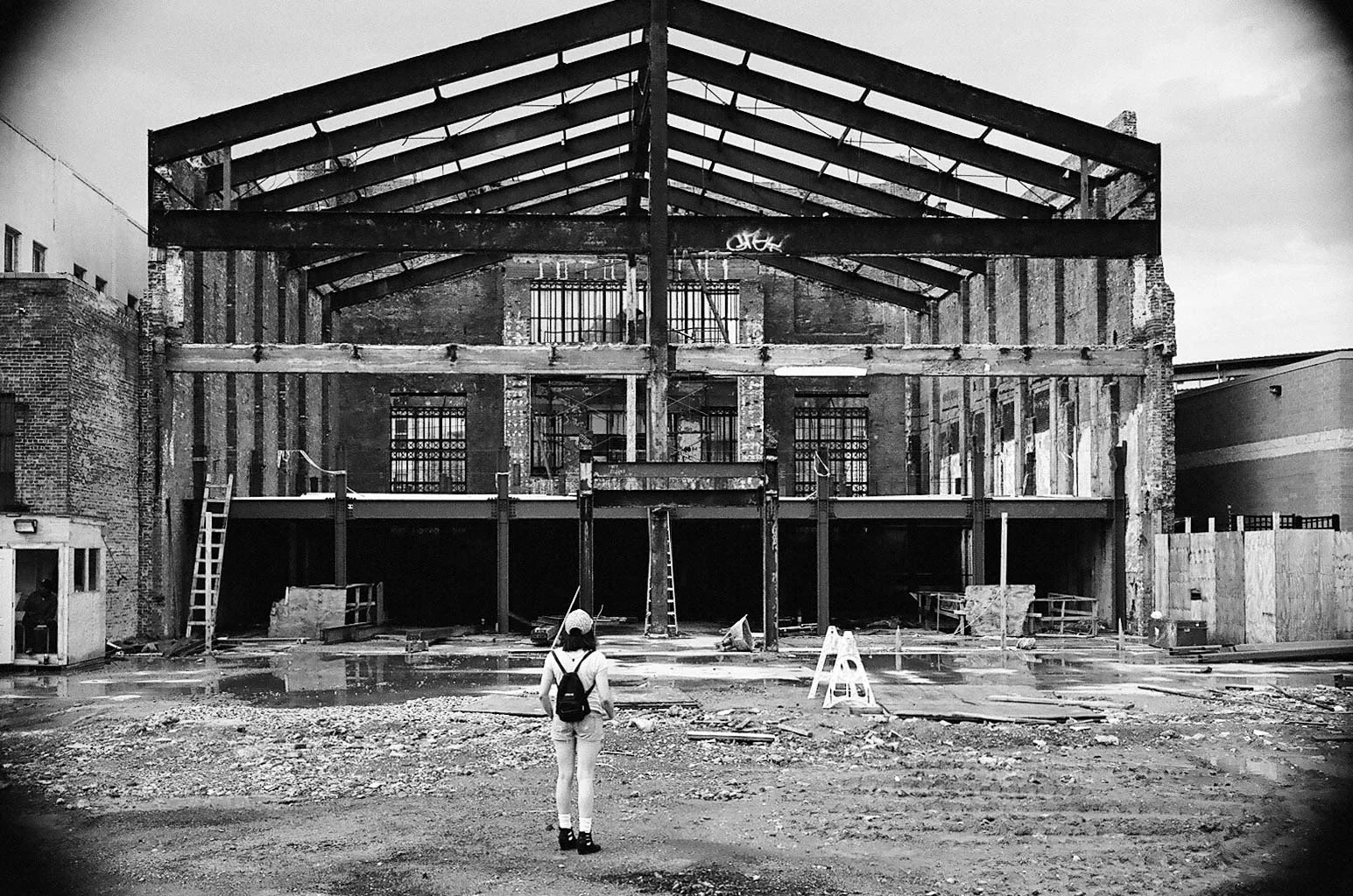 A woman with a backpack stands in front of a partially constructed building with exposed wooden beams and scaffolding, in black and white.