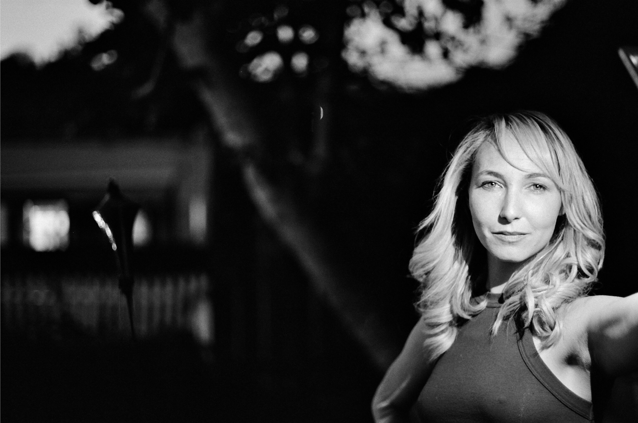 Black and white photo of a woman with long blonde hair smiling outdoors at night, with blurred lights and trees in the background.