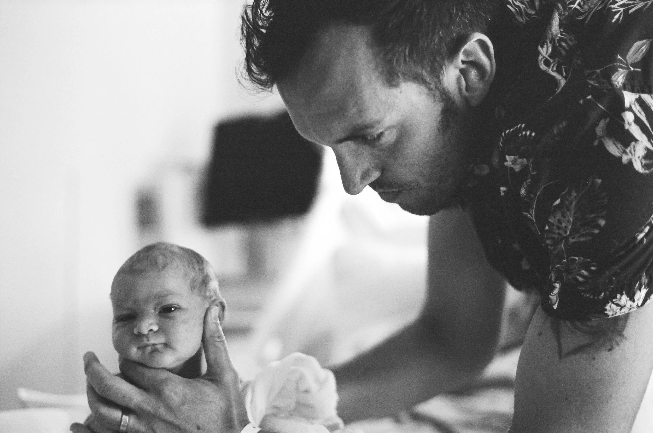 A black-and-white photo of a man with curly hair and a beard, smiling as he looks down at a baby. The baby has short hair, is smiling, and has a hand near his face. The man is wearing a patterned shirt, and the background is plain and out of focus.