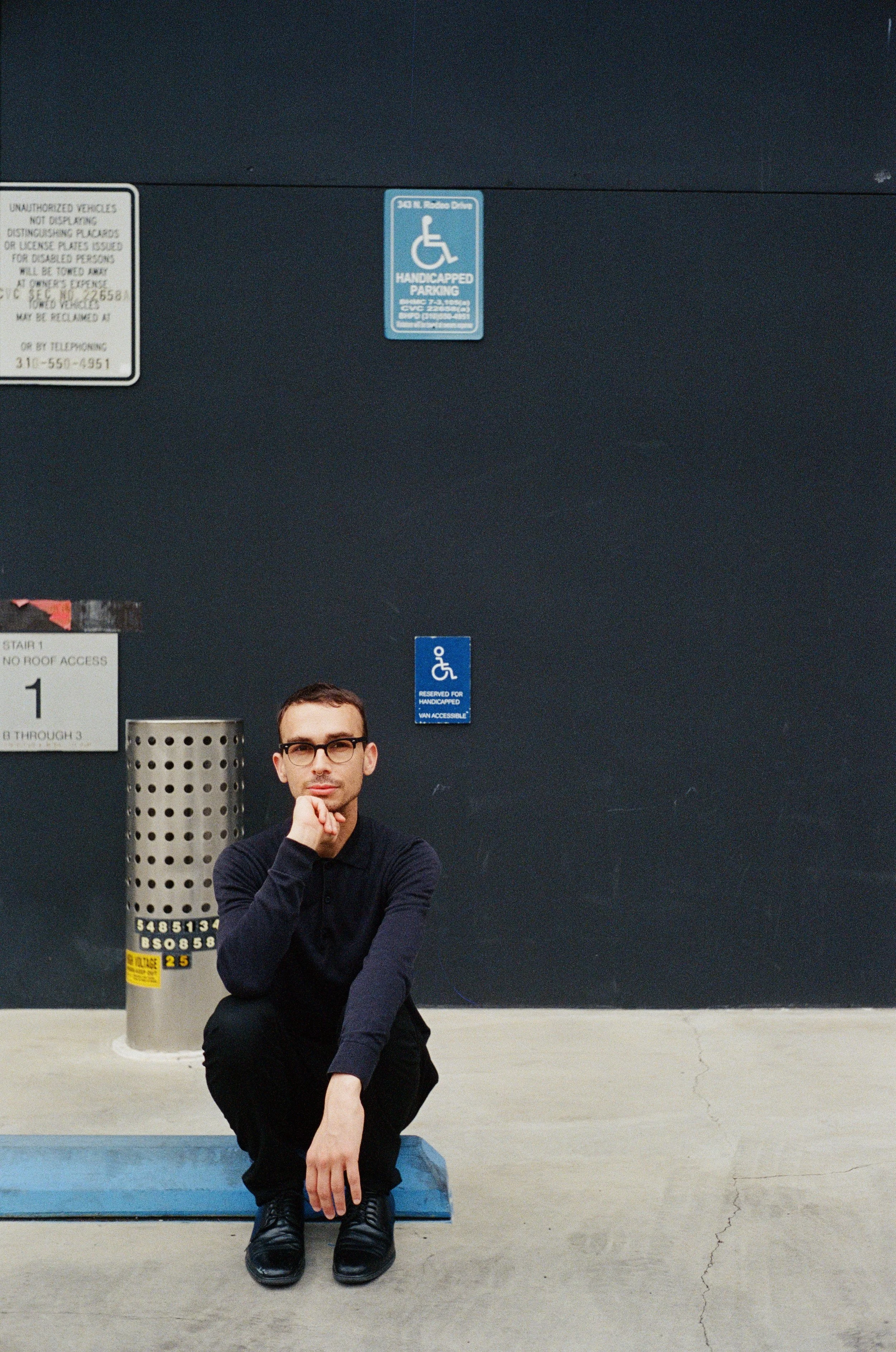 A man in glasses and dark clothing crouches down in front of a parking lot wall with parking signs, a meter, and a blue parking block, sitting on the curb with one hand on his knee and the other touching his face.