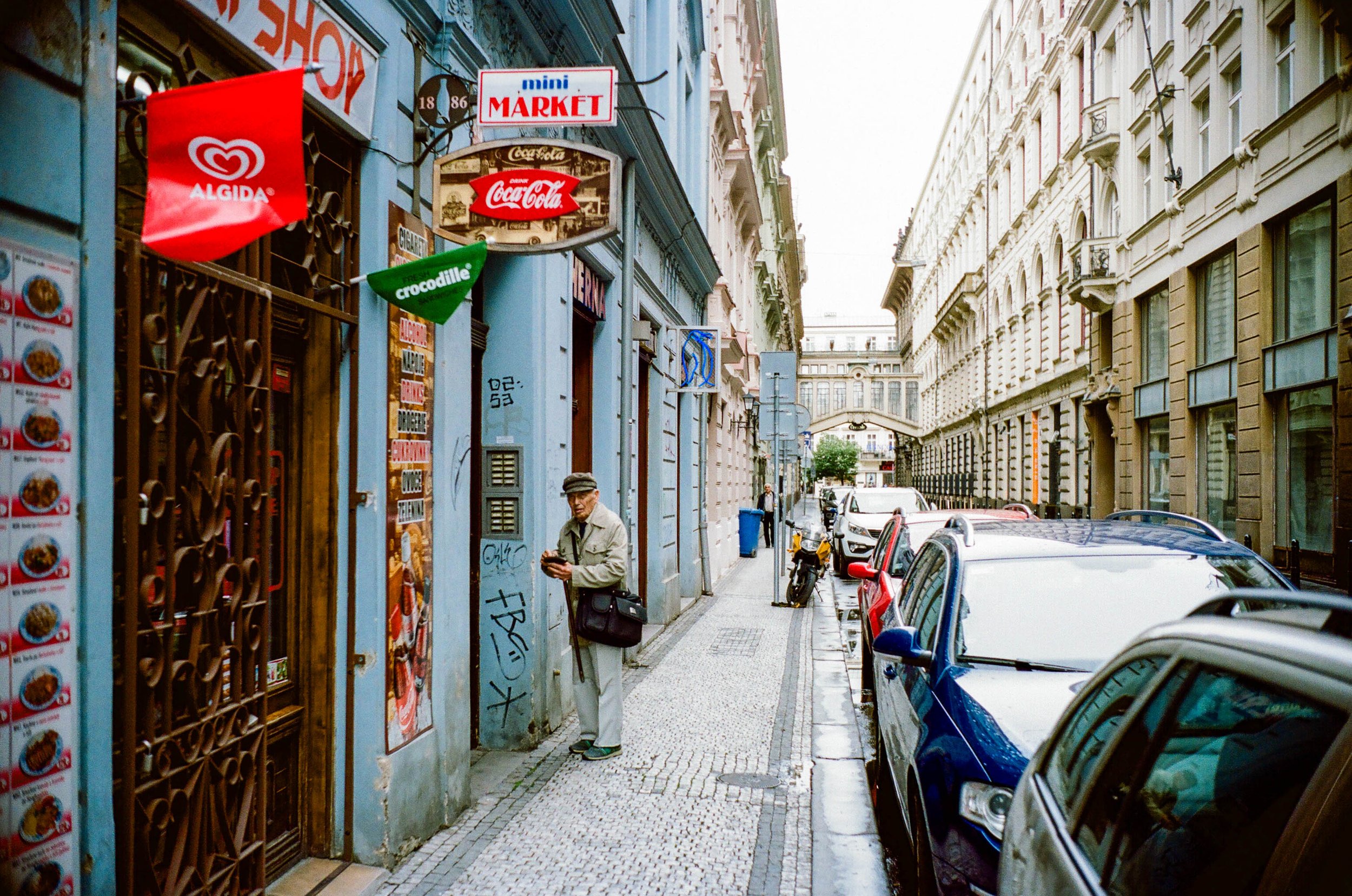 Street scene in an urban area with parked cars, pedestrians, colorful buildings, and signs for small stores including a mini market, Coca-Cola, and Algida ice cream.