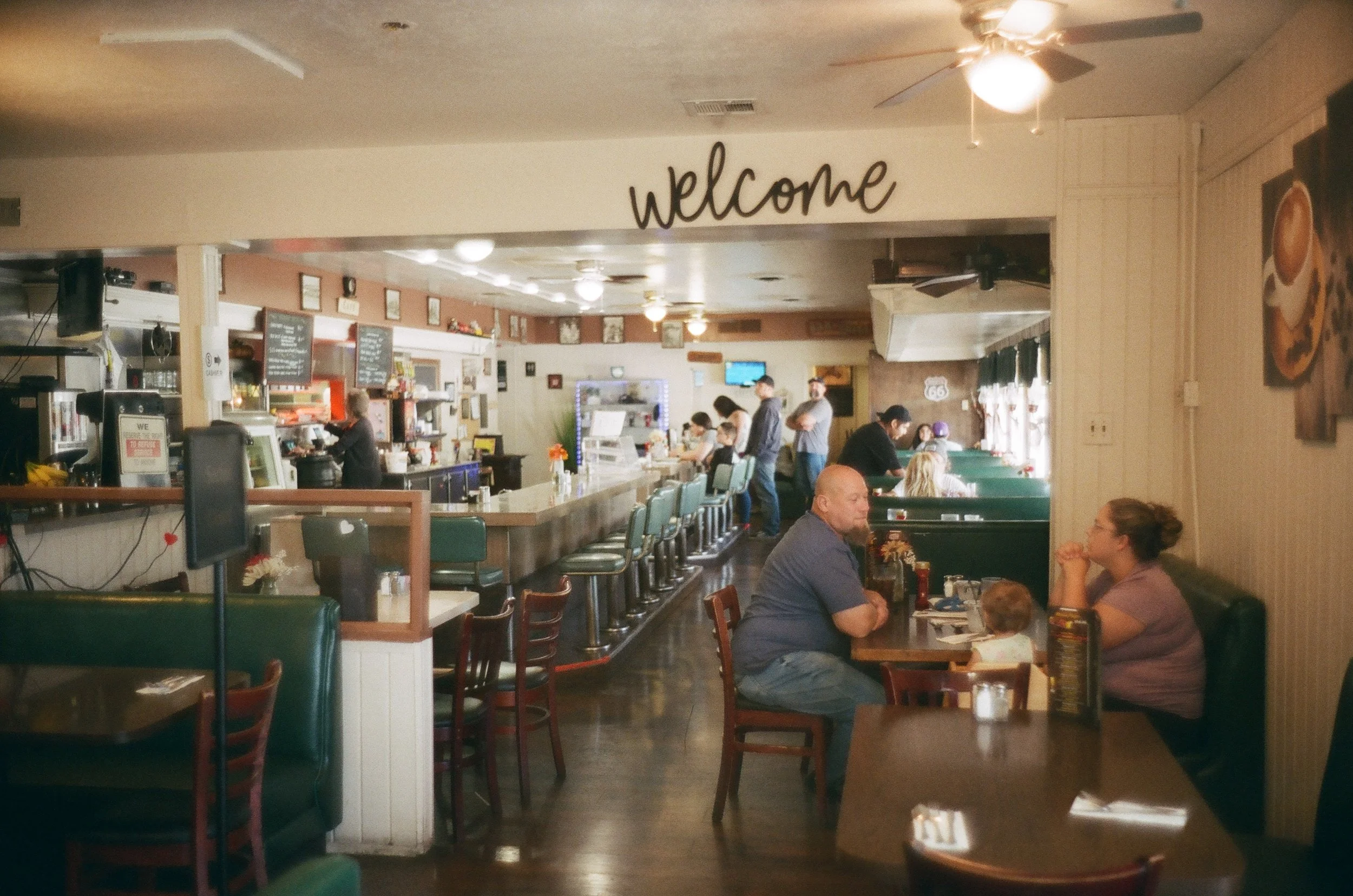 Interior of a restaurant with a bar area, several tables, and chairs, some people dining and standing near the counter. A "welcome" sign is visible above the entrance to the kitchen or service area. The Original Saugus Cafe