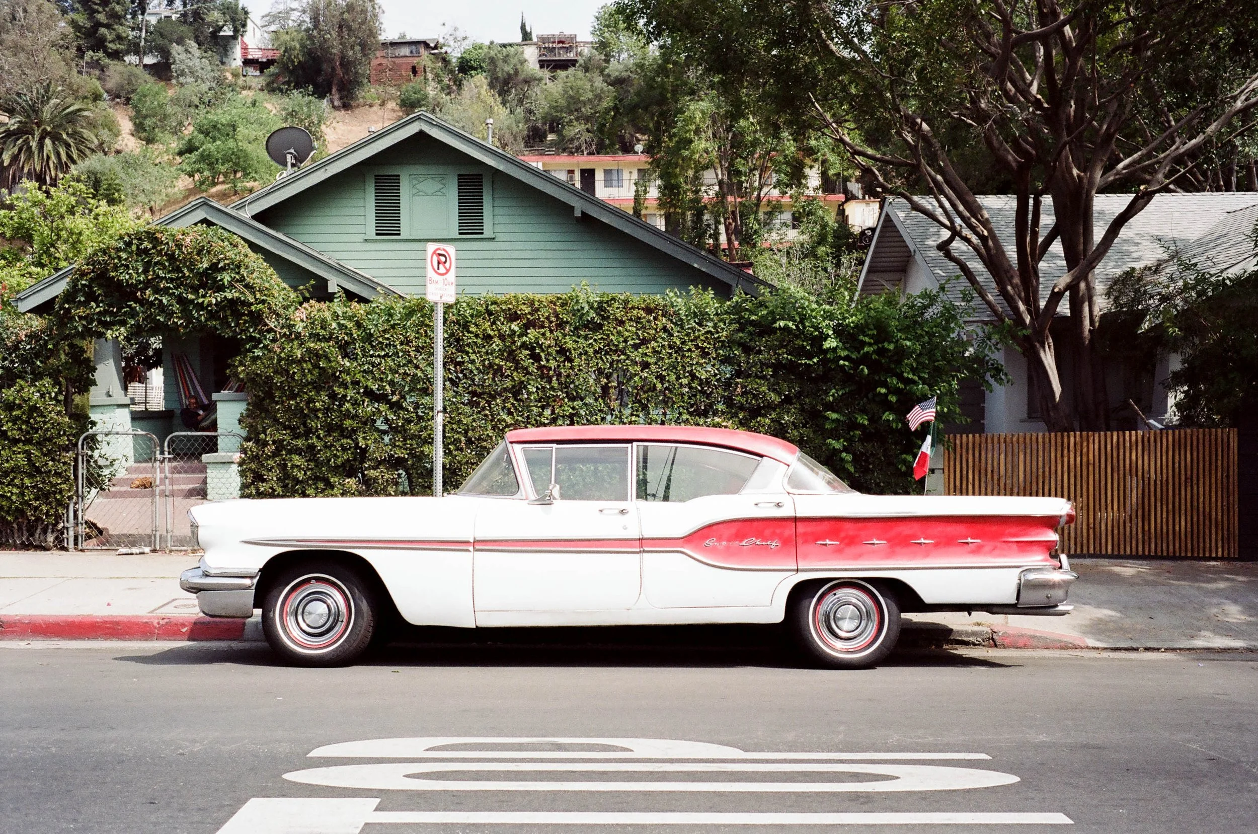 A white classic car parked on the street in front of a hedge and a house with a green roof. There is a no parking sign on a pole behind the car, and a small American flag near the fence in the background. The scene is set in a suburban neighborhood w