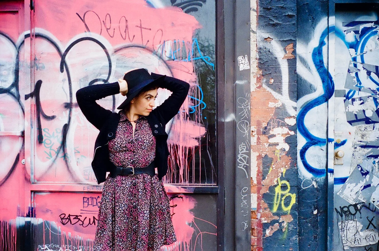 A woman in a floral dress and black jacket holding a black hat, standing in front of a graffiti-covered pink and black wall.