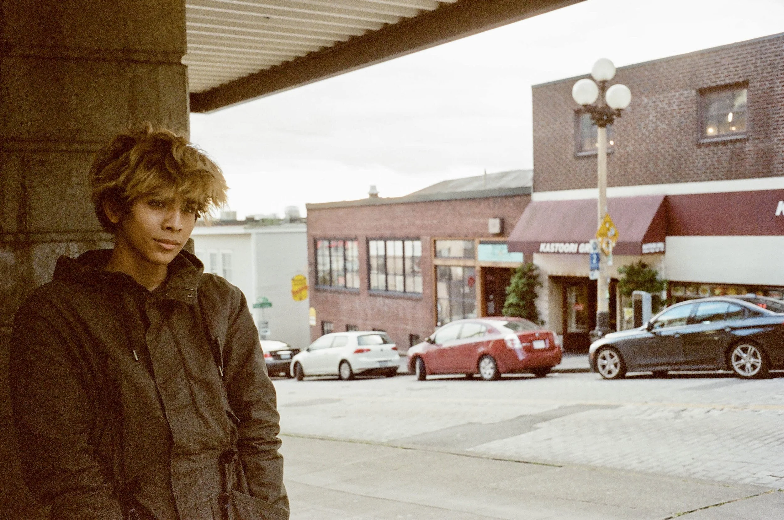 A young person with short, curly hair standing outdoors near a building, with a parking lot and brick buildings in the background on a cloudy day.