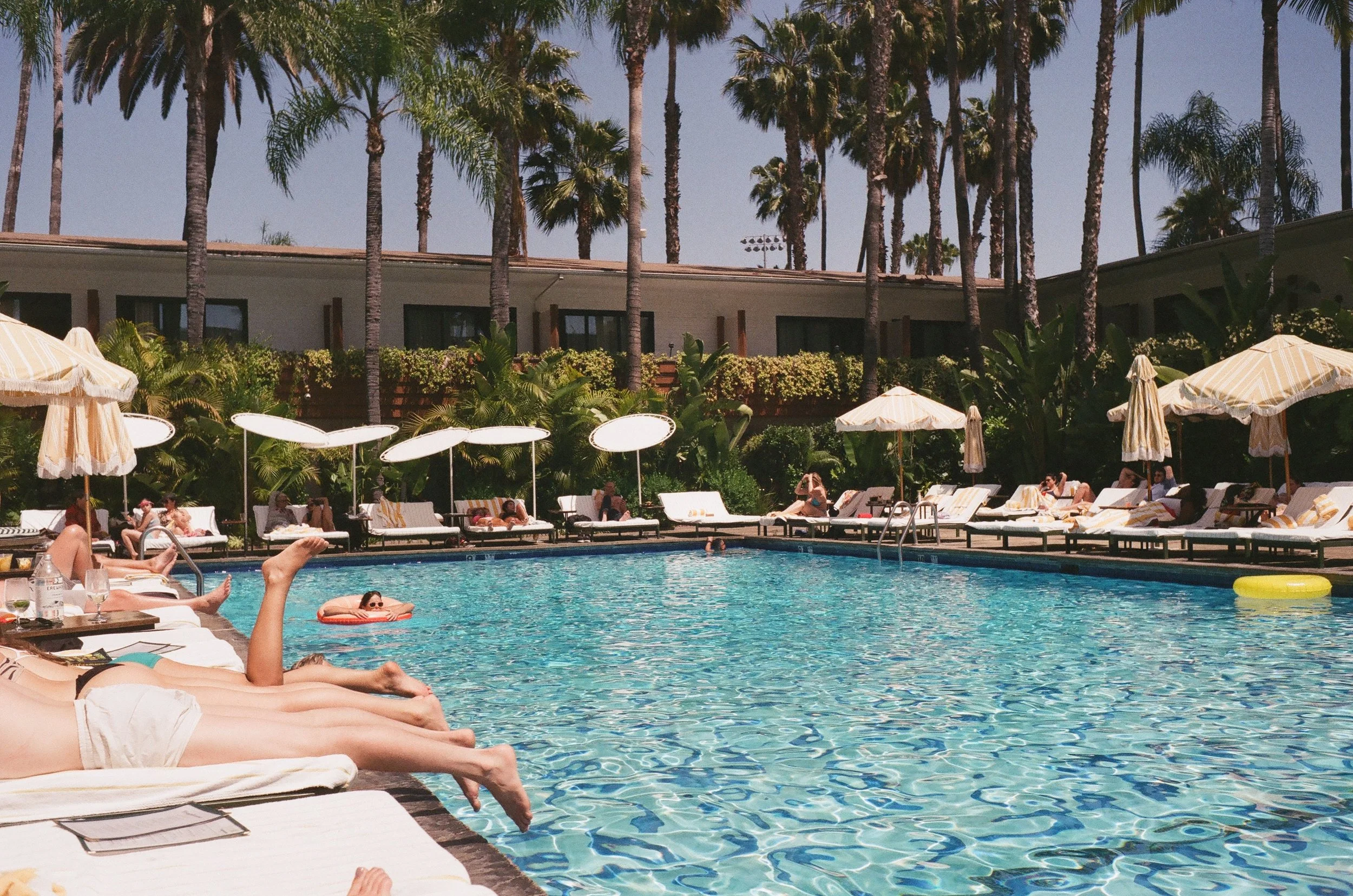 A swimming pool area with people lounging on chairs under umbrellas, some in the pool, with palm trees and a building in the background on a sunny day.
