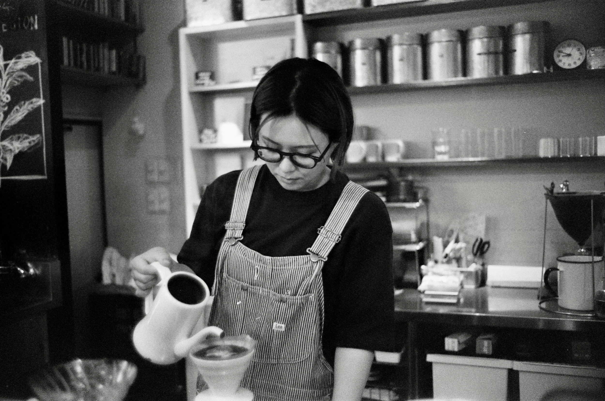 A woman wearing glasses and an apron pouring a liquid into a container in a kitchen or café setting; Tokyo