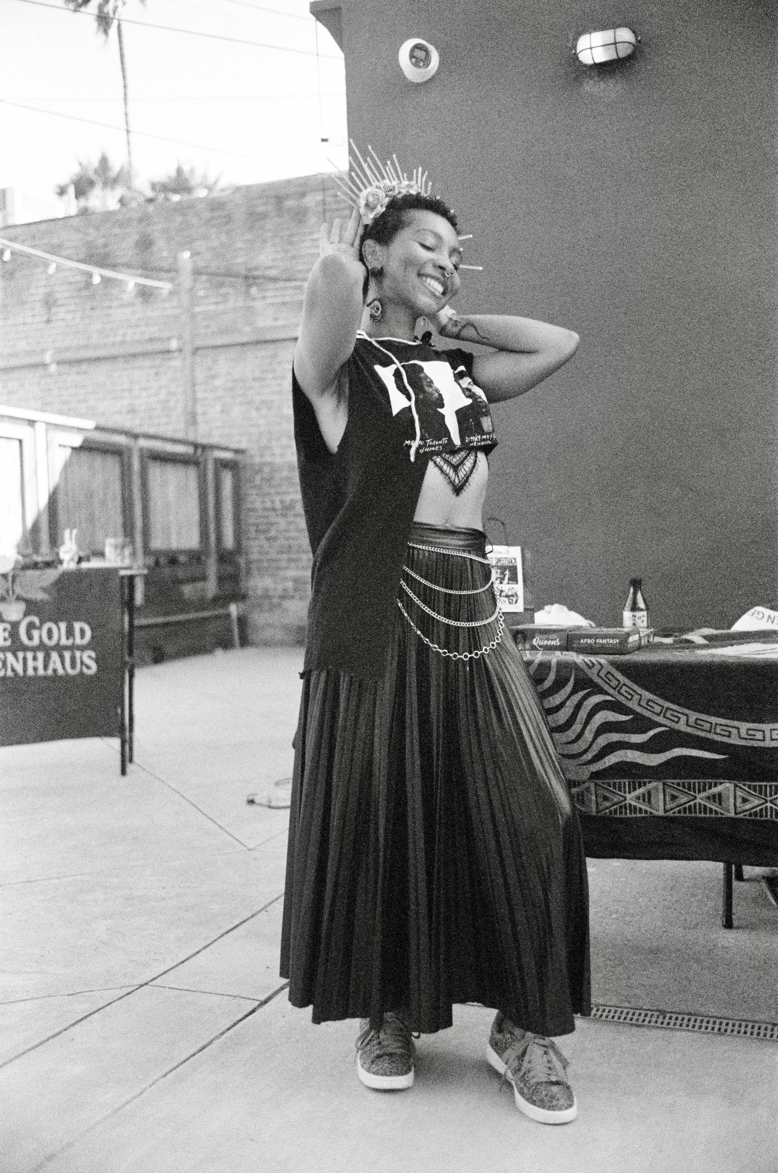 A young woman with short hair smiling and wearing a crown, black graphic crop top, long pleated skirt with chains, and sneakers, standing indoors with a table and decoration visible in the background.