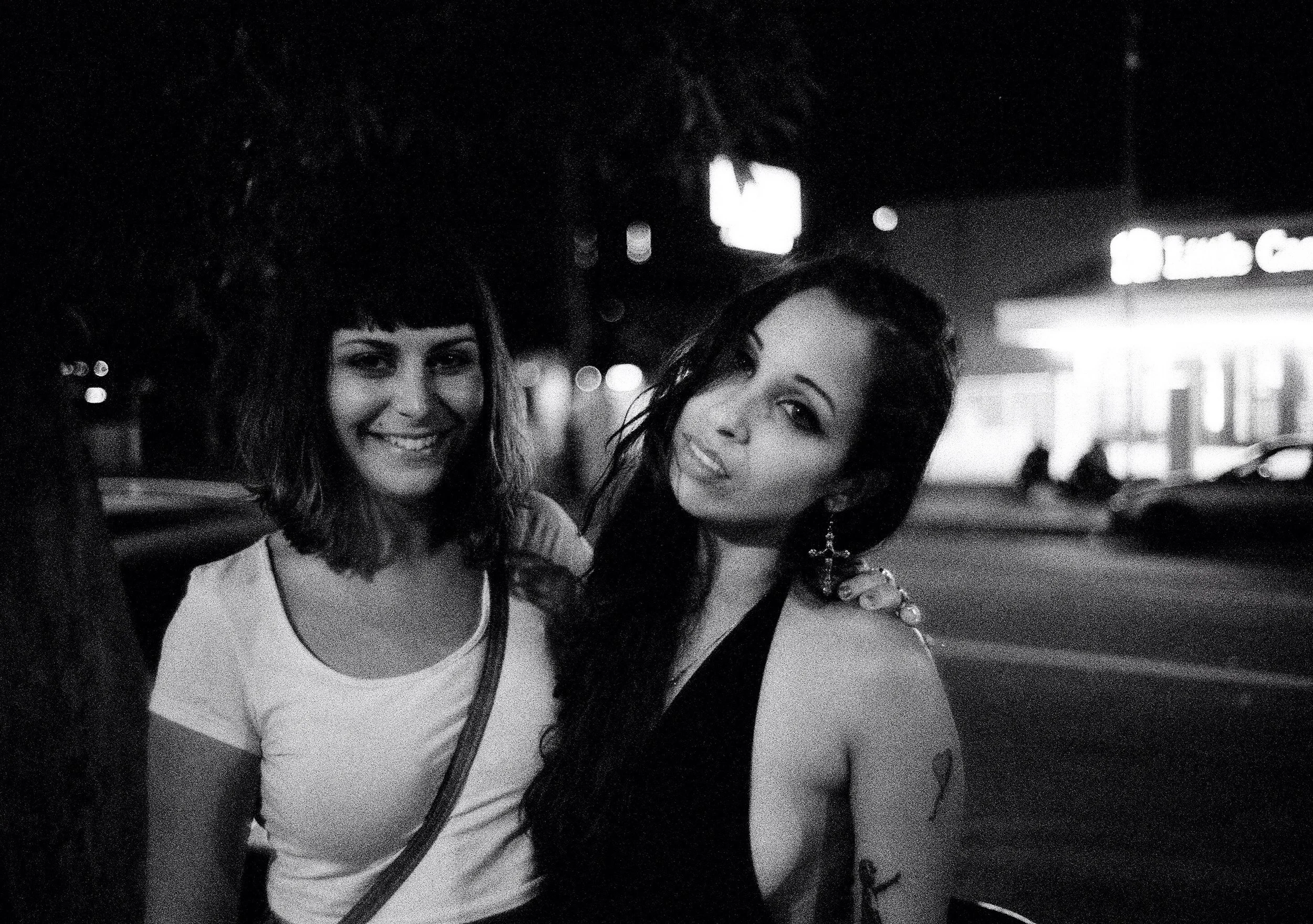 Two women smiling together in a nighttime city setting, with blurred background lights.