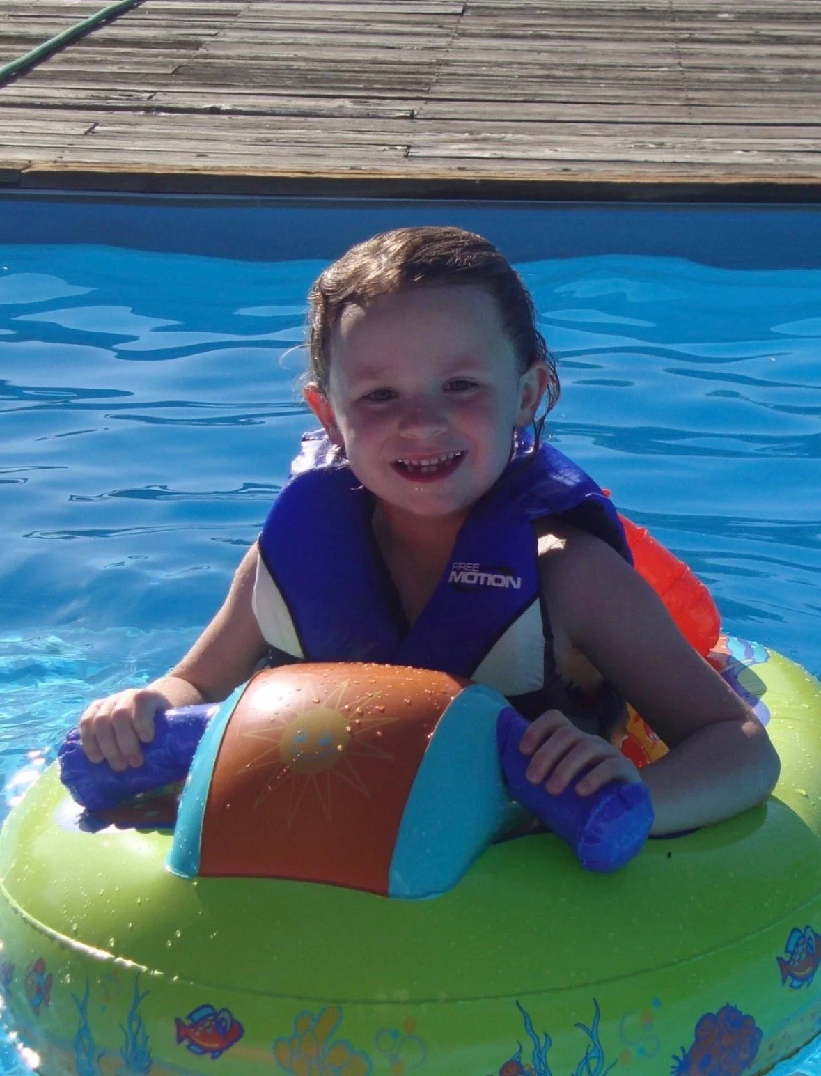 A young child smiling in a swimming pool, wearing a life vest and holding onto a colorful inflatable ring with a sea creature design.