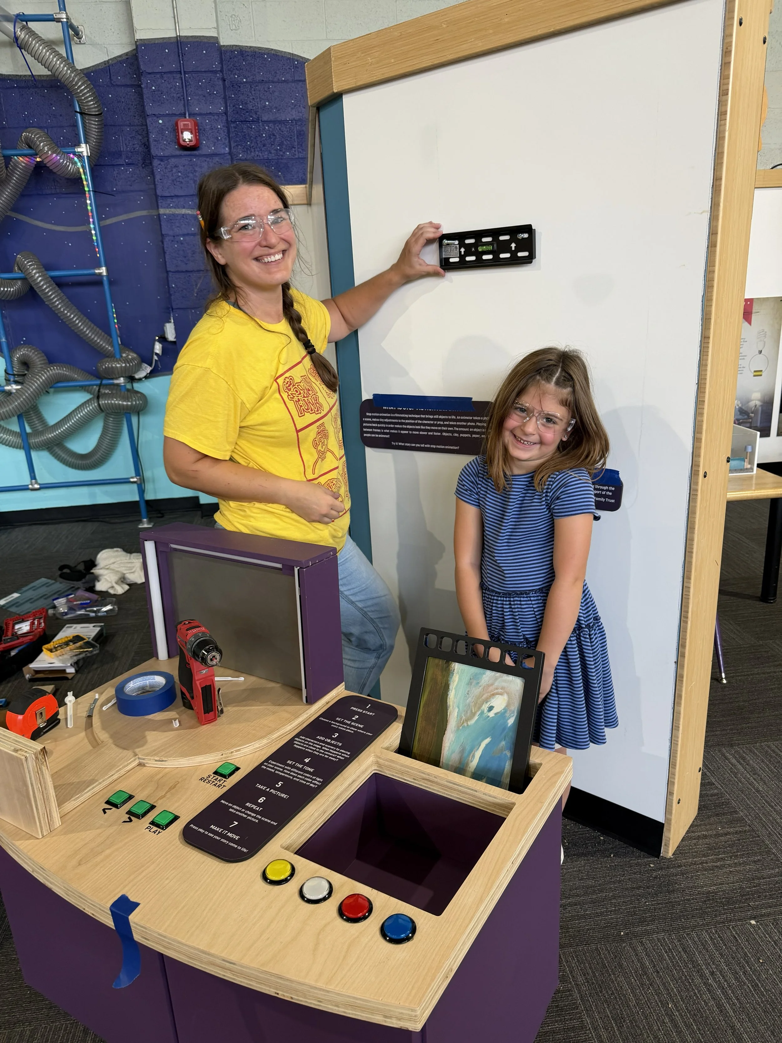 A woman and a girl are smiling and holding a game controller at a tabletop game setup featuring a monitor, buttons, instruction card, and various tools. The setup is in a room with tools and a colorful backdrop.