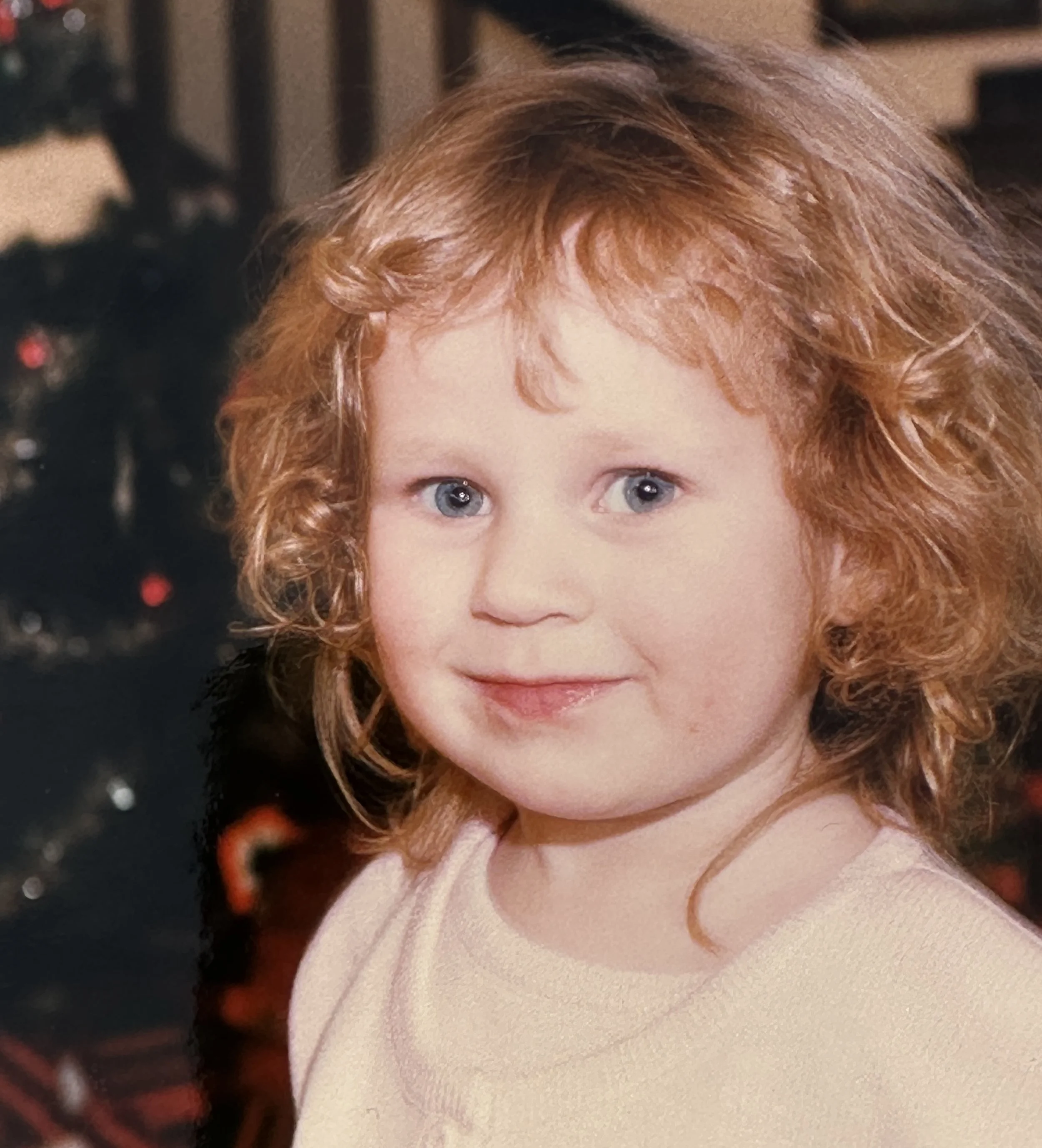 Close-up of a young girl's face with blue eyes and curly red hair, wearing a soft pink sweater, in front of a blurred Christmas tree with dark green branches and red ornaments.