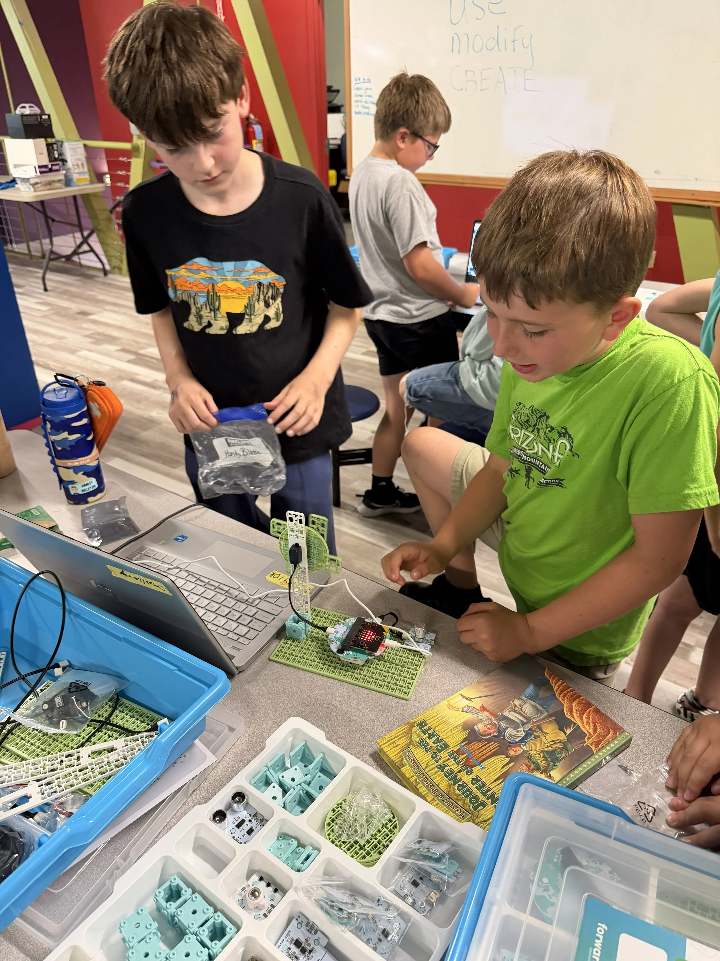 Children working on an electronics project at a table filled with various electronic components, tools, and kits in a classroom setting.