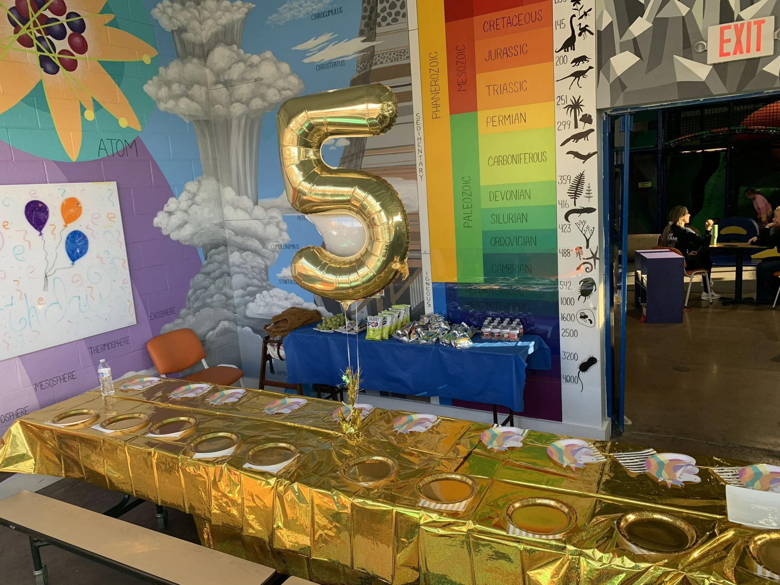 Party table decorated with a gold tablecloth, paper napkins, and paper plates, with a golden balloon shaped like the number 5 attached to a stand. The background has a colorful education mural with clouds, volcanoes, and a chart of geological periods.