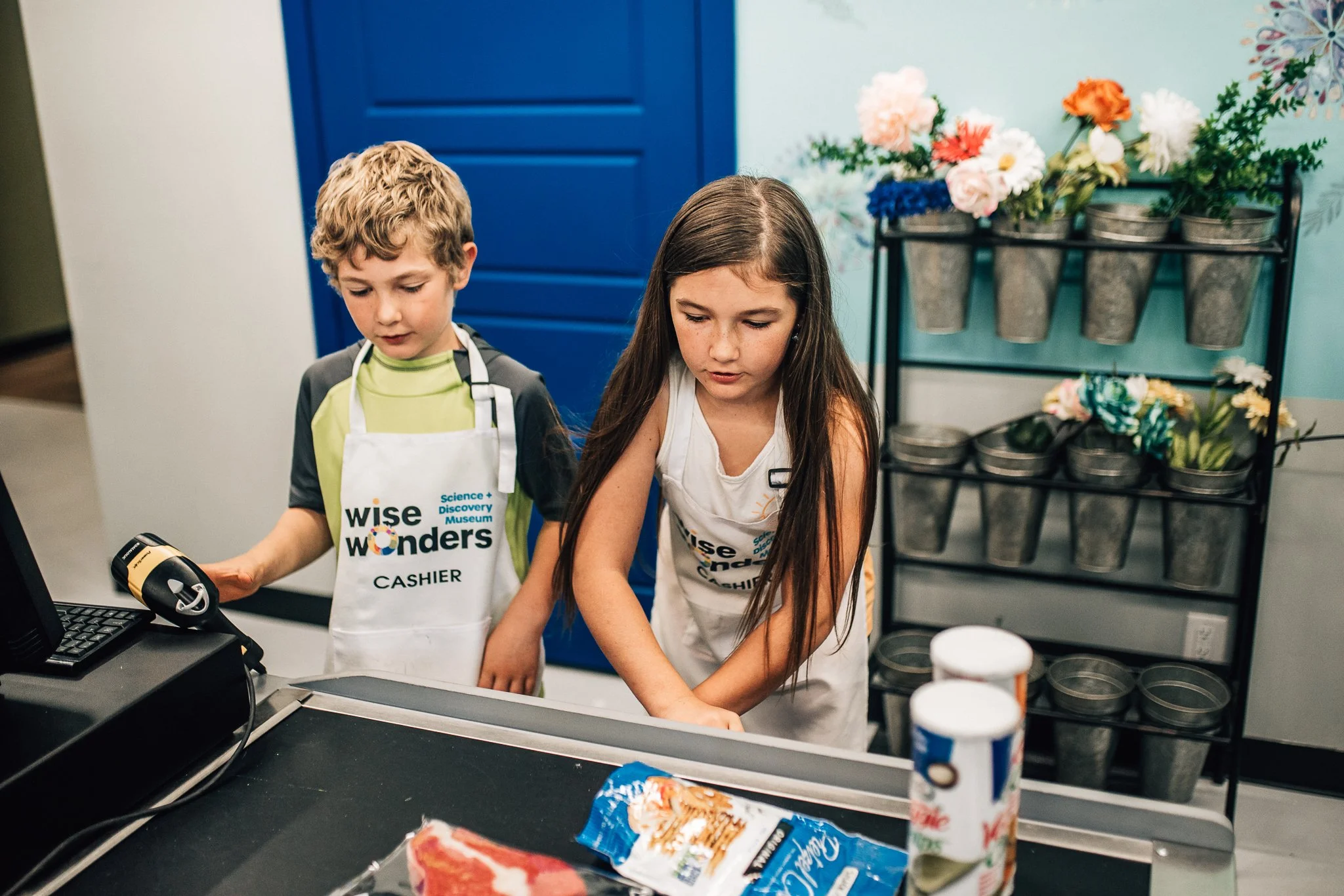 Two children at a checkout counter in a store, one of whom is a boy wearing an apron that reads 'wise wonders' and 'cashier,' the other a girl with long dark hair. The background shows a blue door, plant pots, and colorful flowers.