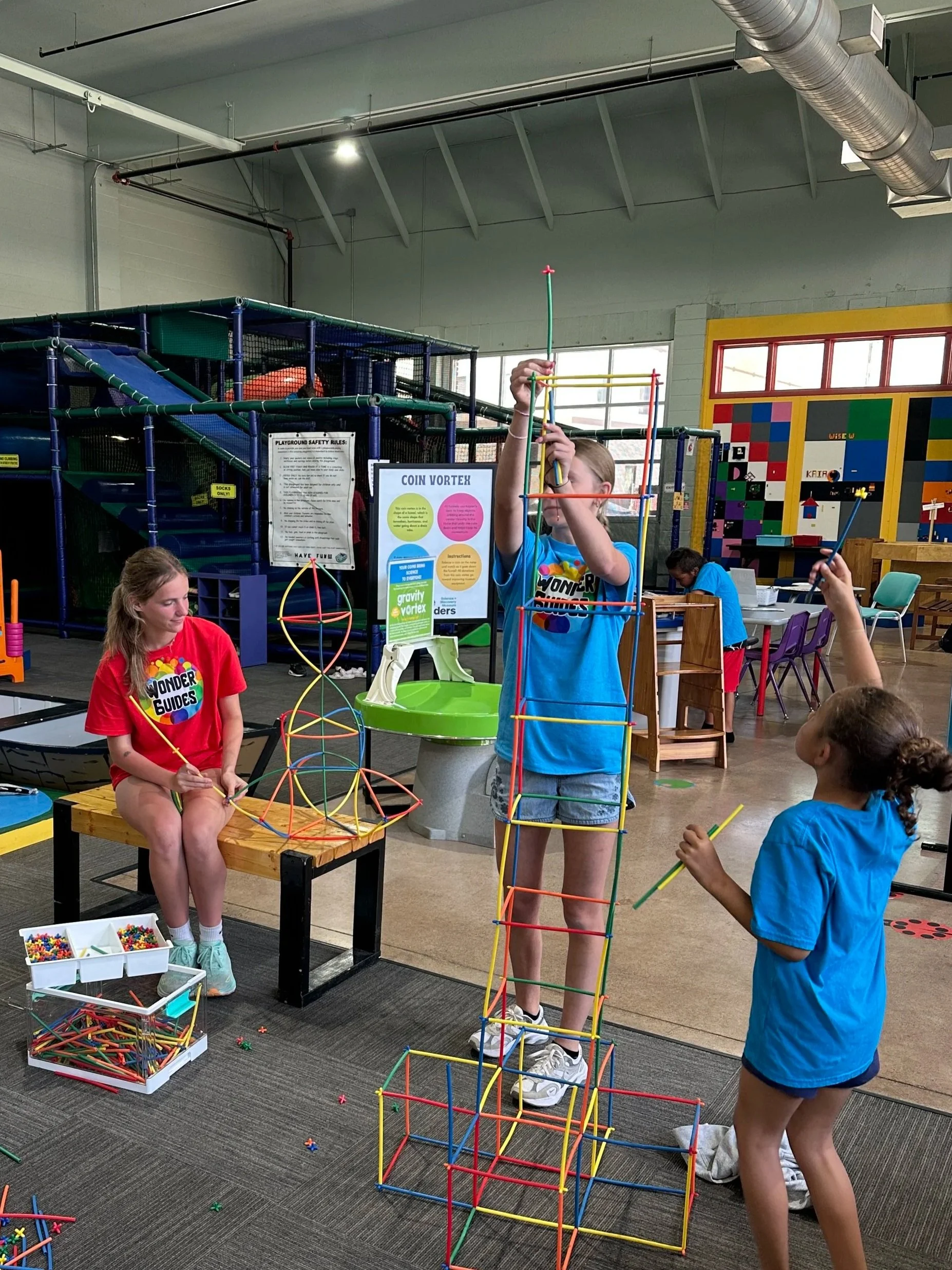 Children building a large structure with colorful plastic sticks in an indoor play area.