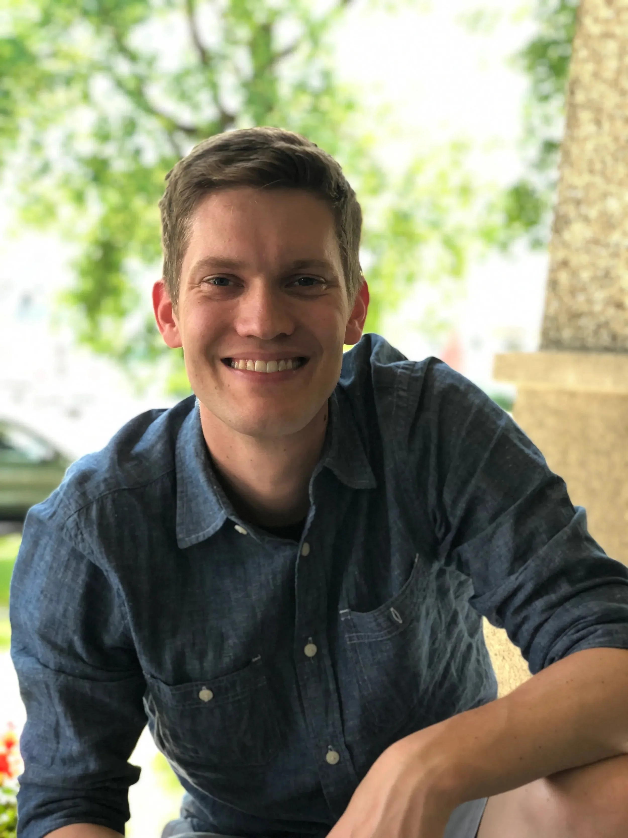 A smiling young man with brown hair wearing a dark blue and black plaid shirt, sitting outdoors with green trees in the background.