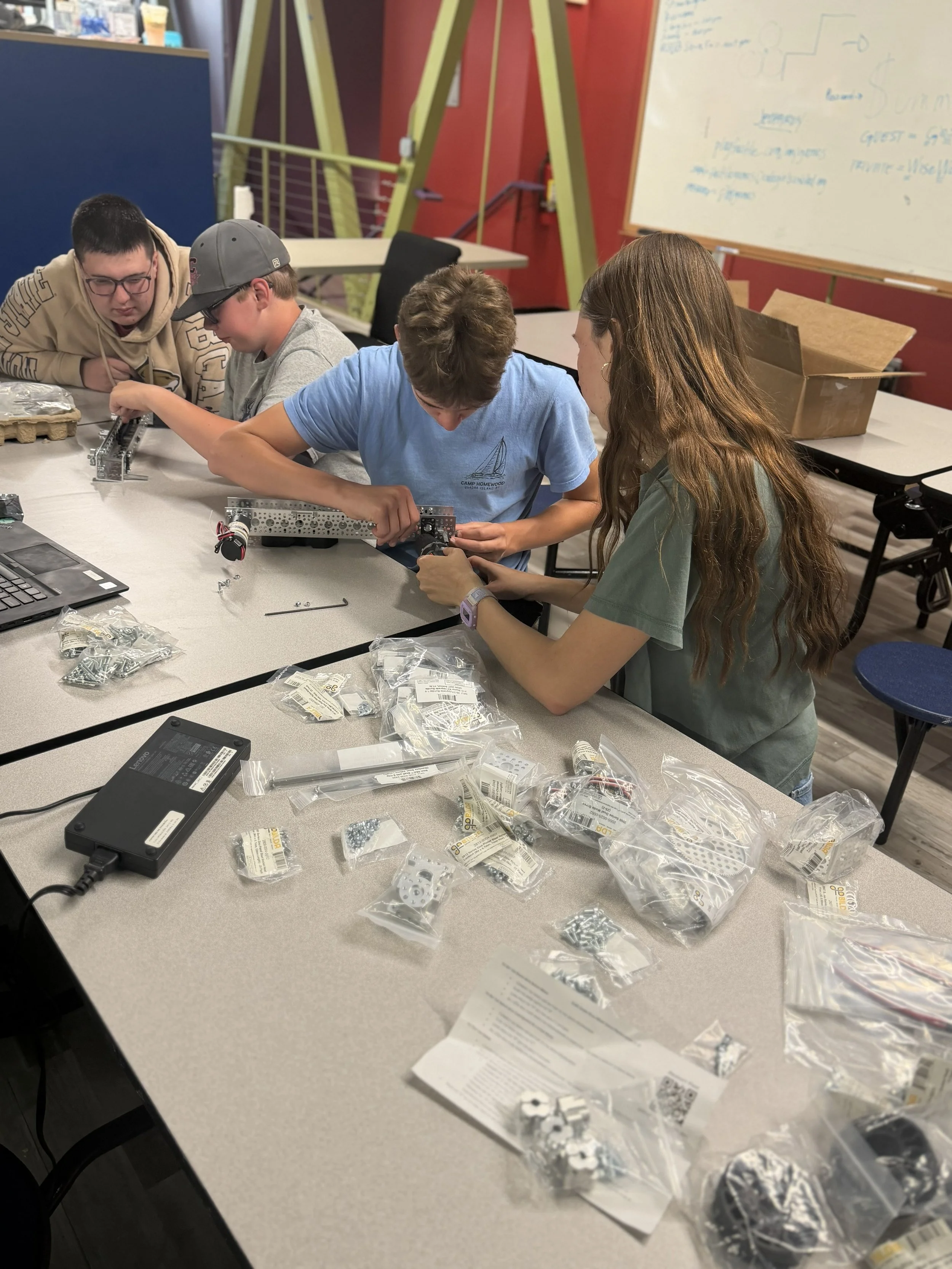 Four young people working together on a robotics project at a table covered with parts, tools, and instruction sheets. In the background, there is a whiteboard with notes and diagrams, and a box of additional parts.