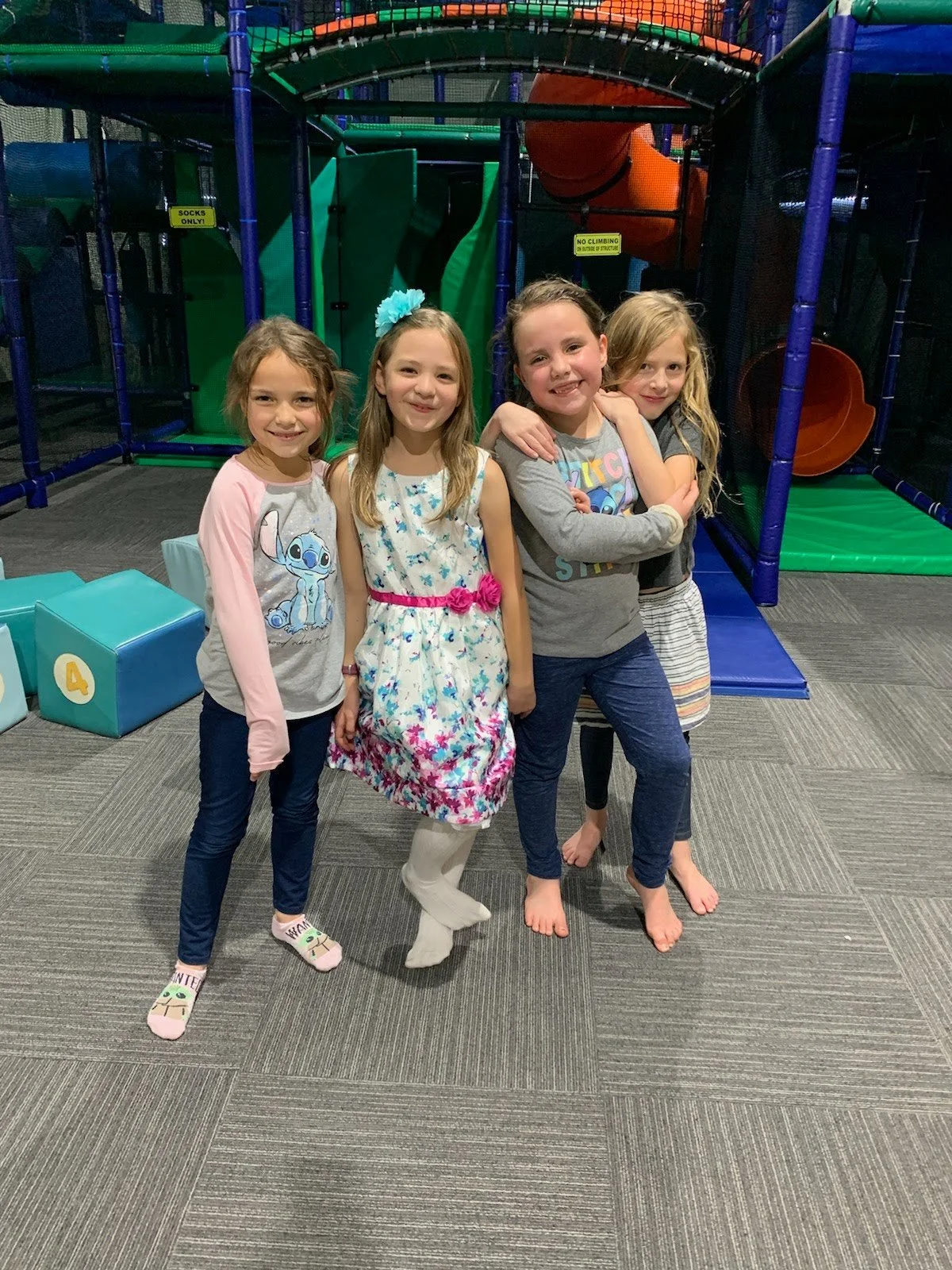 Four young girls standing in front of an indoor play structure at a playground or indoor amusement center, smiling at the camera.