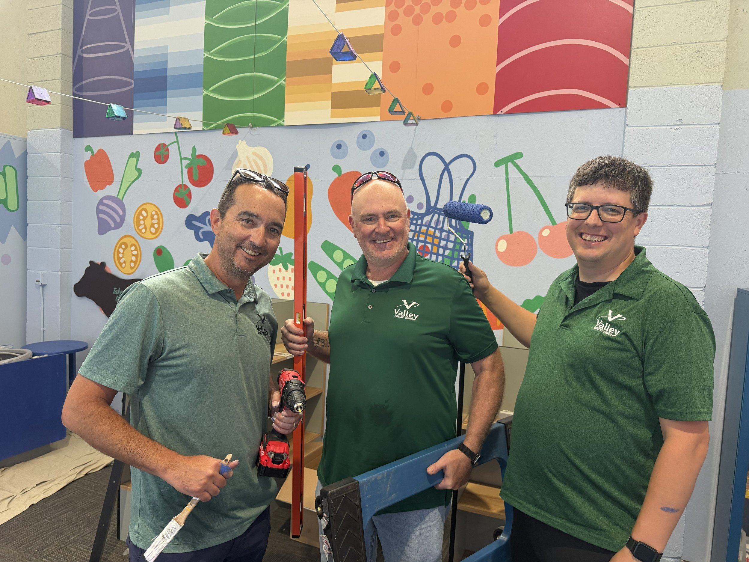 Three men wearing green Valley Credit Union shirts smiling at the camera inside a colorful room with a painted wall featuring fruit and vegetable illustrations. The middle man holds a construction level and a power drill, while the others assist with construction or painting tasks.