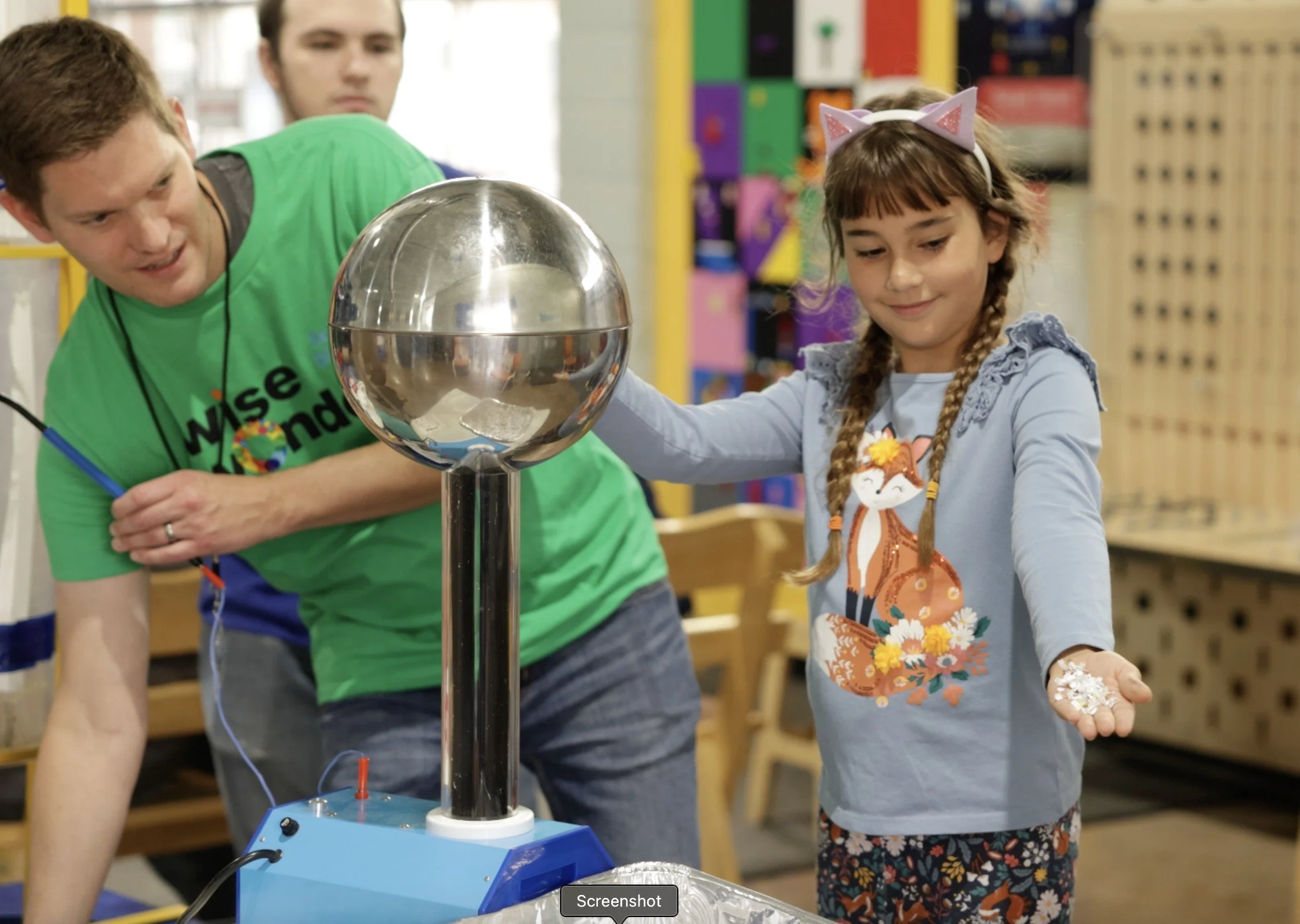 Two children, a boy and a girl, are playing with a science experiment involving a large metal sphere and a Tesla coil in a classroom or science center.
