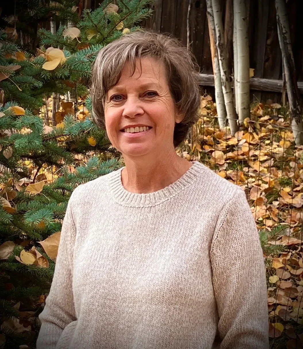 A woman smiling outside during fall, standing in front of a small evergreen tree with yellow leaves and a wooden fence with autumn leaves on the ground.