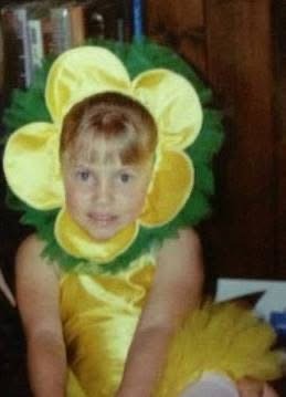 Child dressed in a flower costume, sitting with a wooden background.