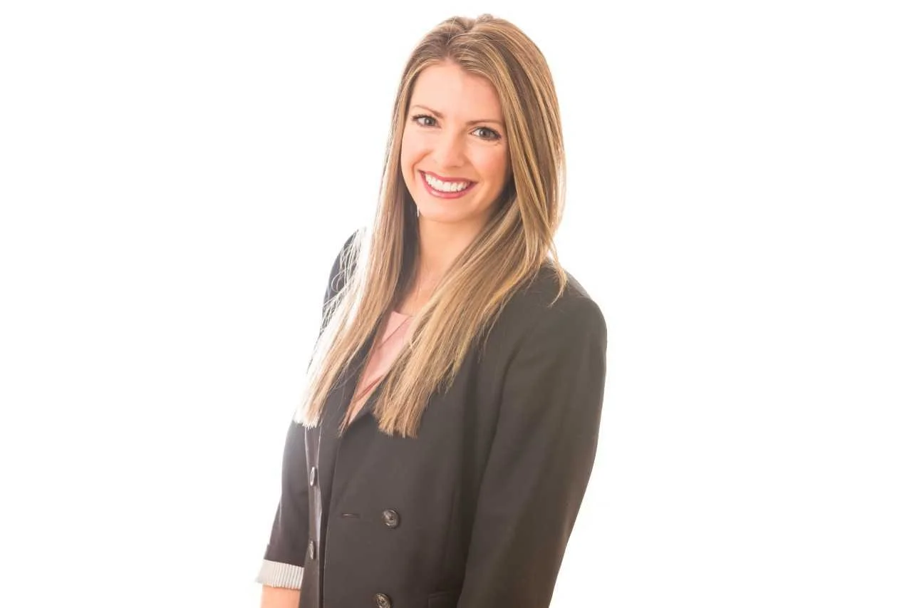 A woman with long blonde hair, smiling, wearing a black blazer over a light pink top, against a white background.