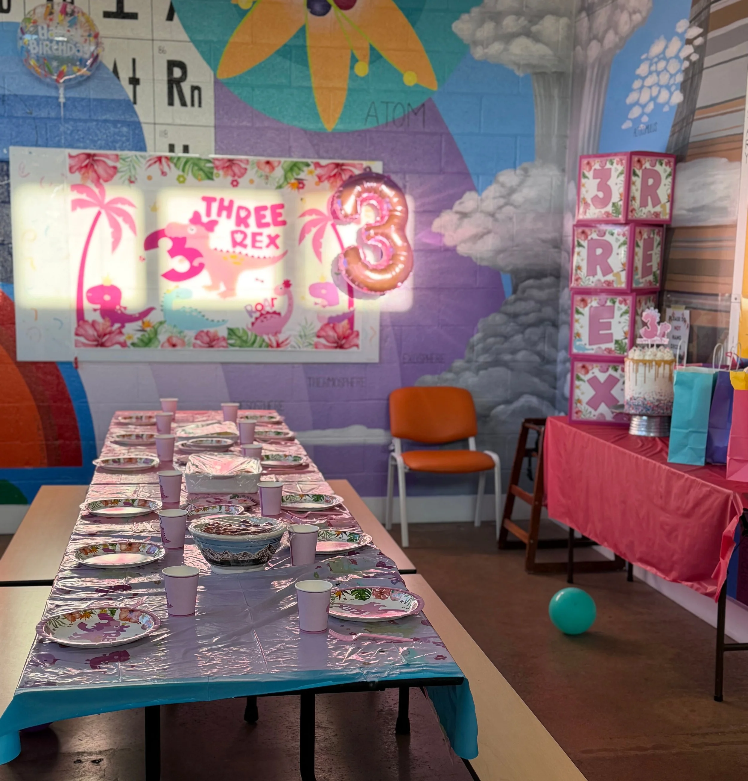 Party table decorated with pink flamingo theme, pink plates, cups, and a balloon shaped like the number 3, with a colorful wall mural in the background featuring cartoons, clouds, and the word "ATOM."