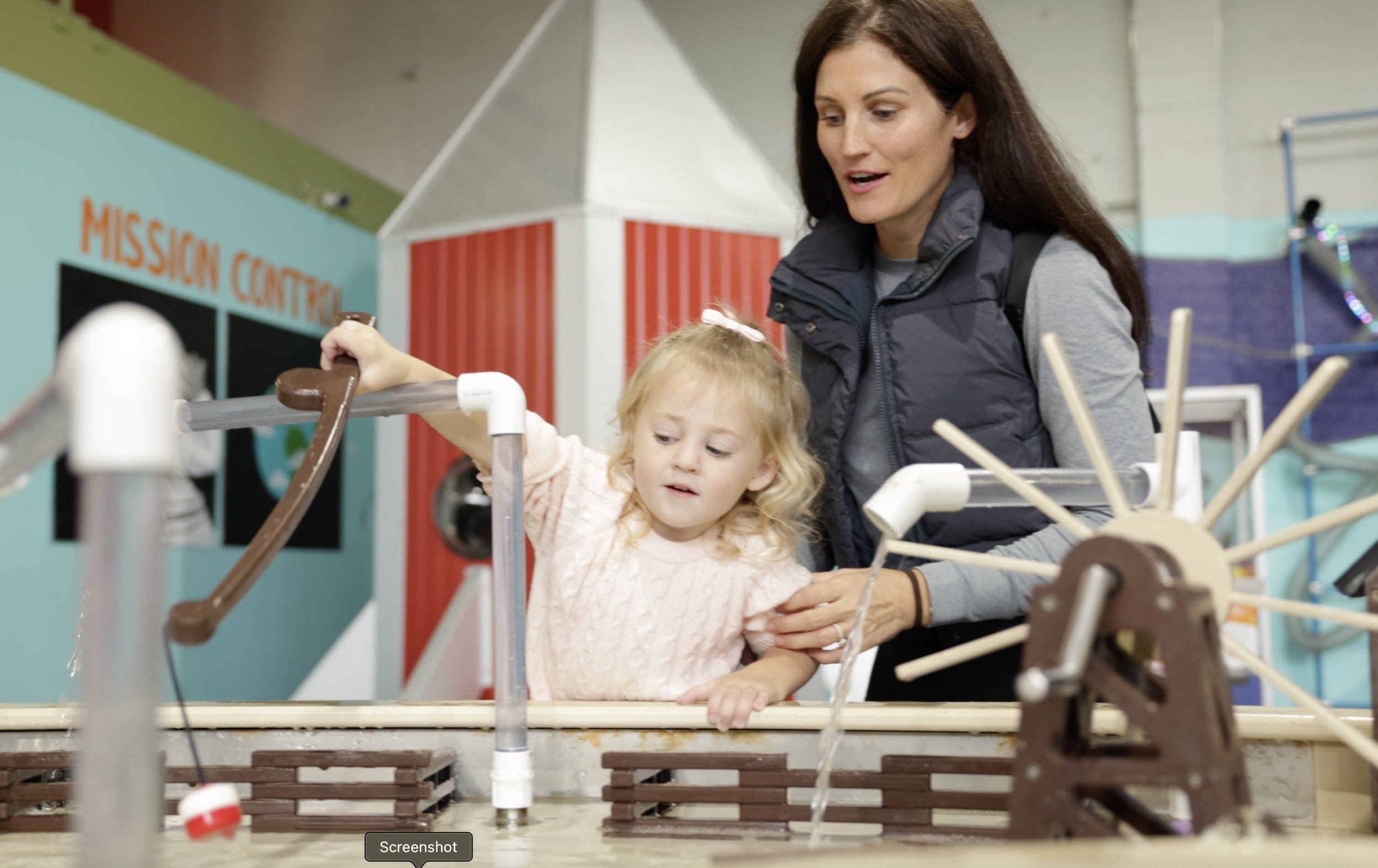 A woman and a young girl standing at a science exhibit involving water and mechanical structures, with a sign reading 'MISSION CENTER' in the background.