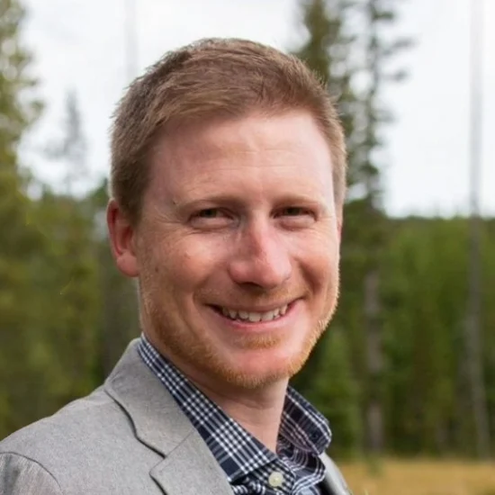 A smiling man with short light brown hair, wearing a gray blazer and checkered shirt, outdoors with trees in the background.