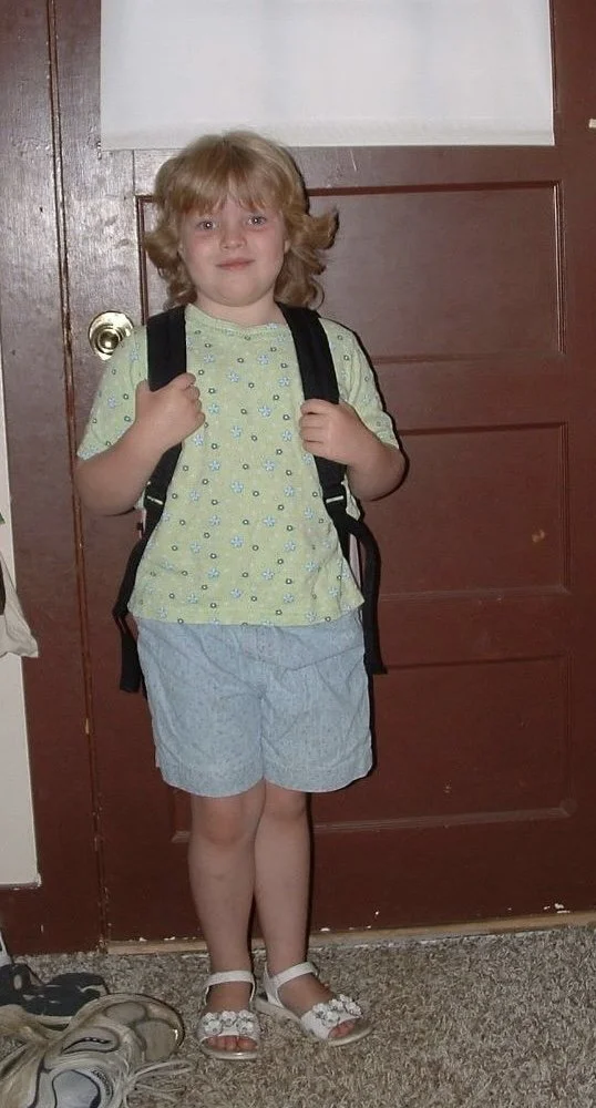Child with blonde curly hair wearing a yellow t-shirt with small blue and purple flower prints, light blue shorts, white sandals with flowers, and a black backpack, standing in front of a brown door.