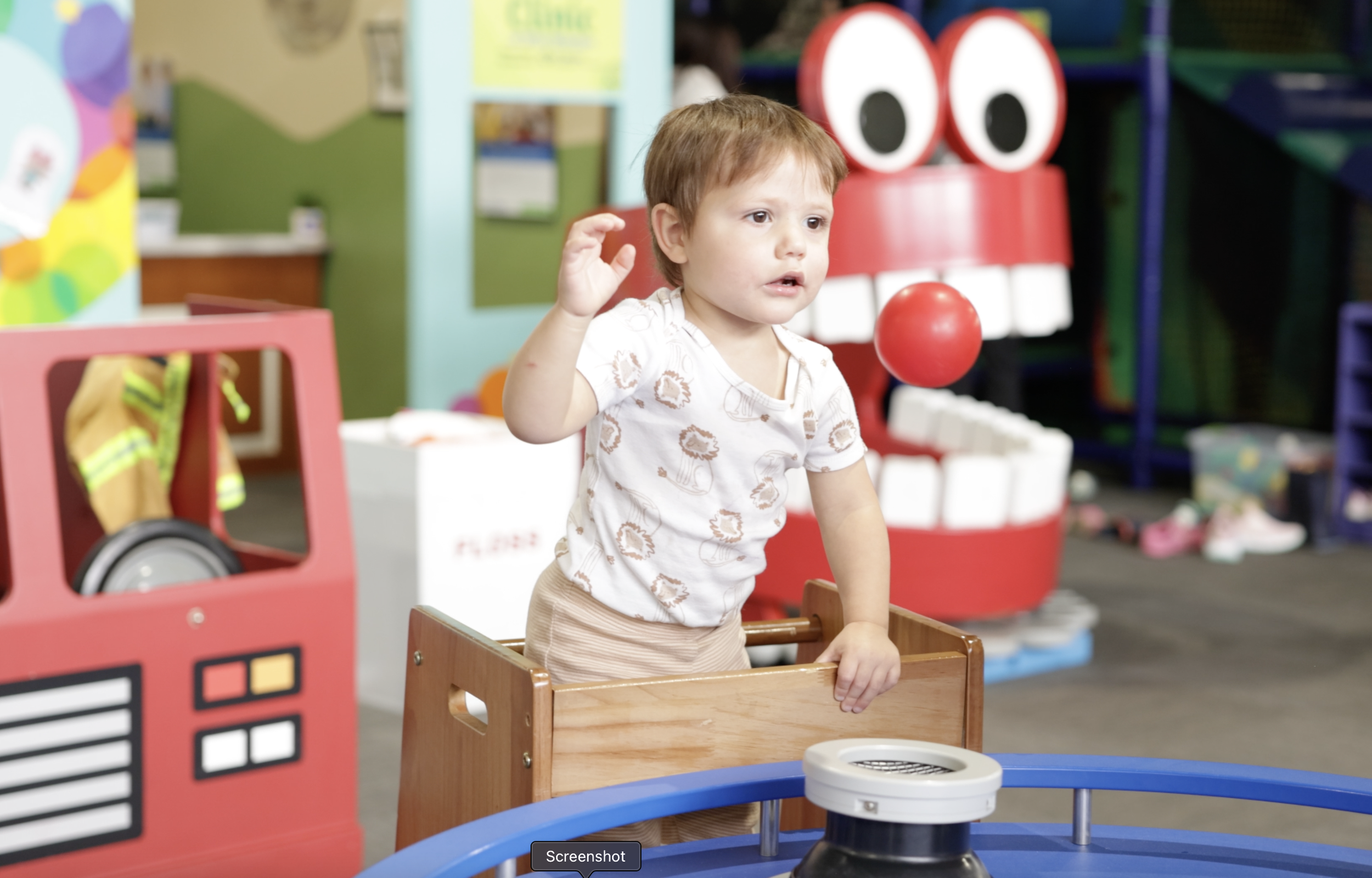 Young boy with brown hair and light skin, raising his right hand, standing inside a play area, likely at a children's museum or play center, with colorful toys and walls visible in the background.
