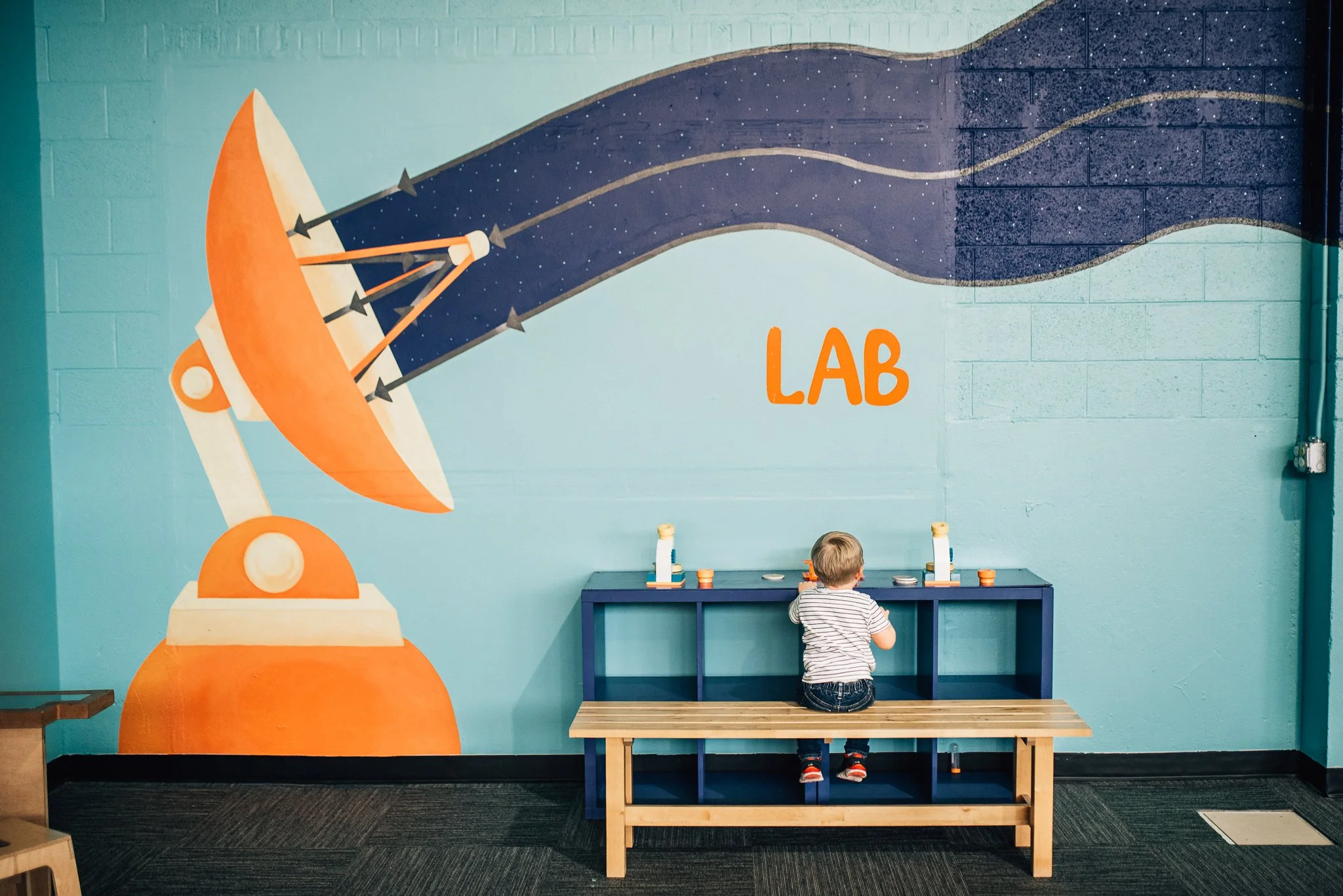 A young boy with blonde hair is kneeling at a blue table in a colorful room. Behind him, there is a large mural on the wall of a rocket ship blasting off into space with a starry background, and the word "LAB" written in orange.