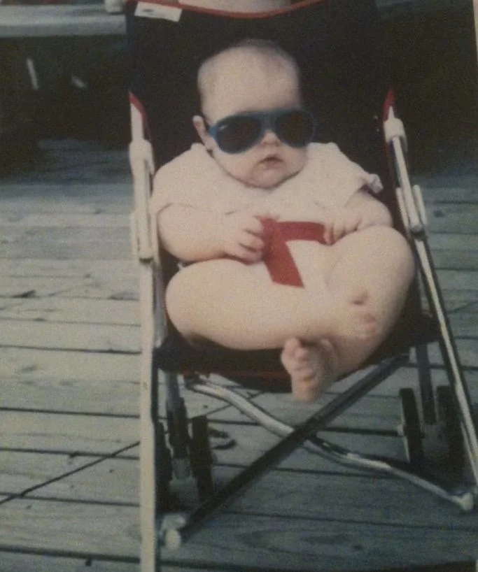A baby seated in a foldable chair on a wooden deck, wearing dark sunglasses and a light-colored outfit, holding a red object, with one leg crossed over the other.