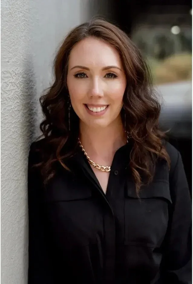 A woman with wavy brown hair, wearing a black top, gold chain necklace, and earrings, smiling and leaning against a textured wall.