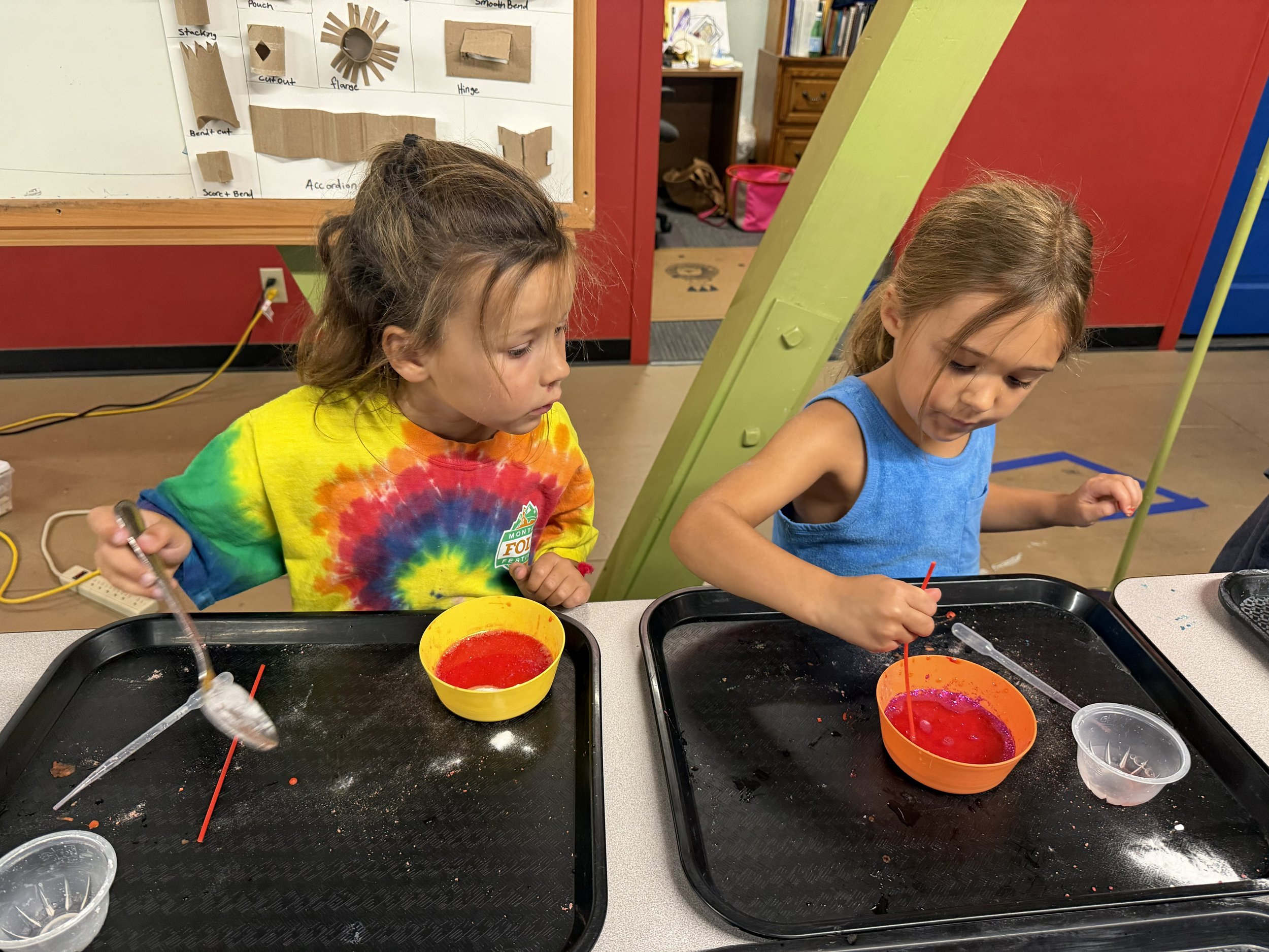 Two young girls sitting at a table engaged in a painting activity. There are trays with red paint, a yellow bowl, and small containers on the table. The background shows a whiteboard with paper crafts and a bookshelf.