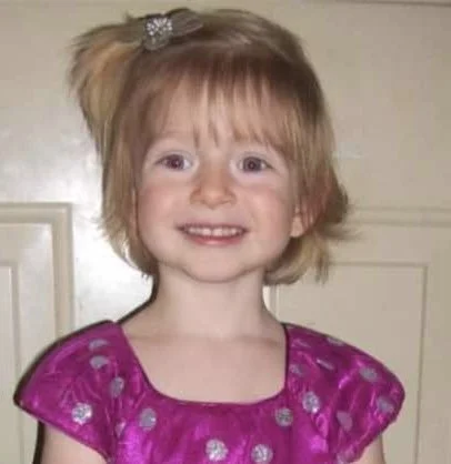 A young girl with blonde hair and a big smile, wearing a pink dress with silver polka dots, and a hair accessory.