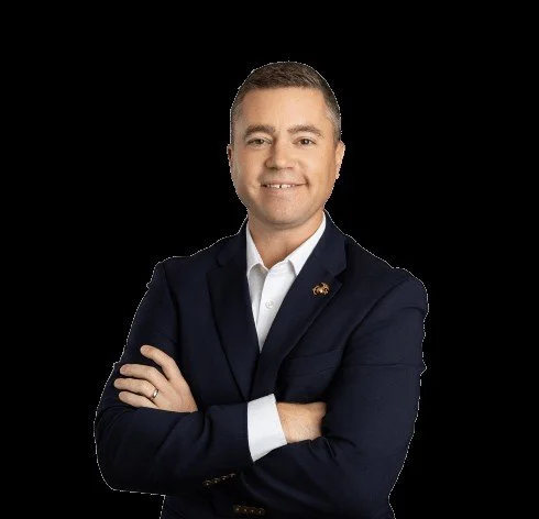 A professional man with short dark hair, wearing a navy blazer over a white shirt, standing with arms crossed and smiling.