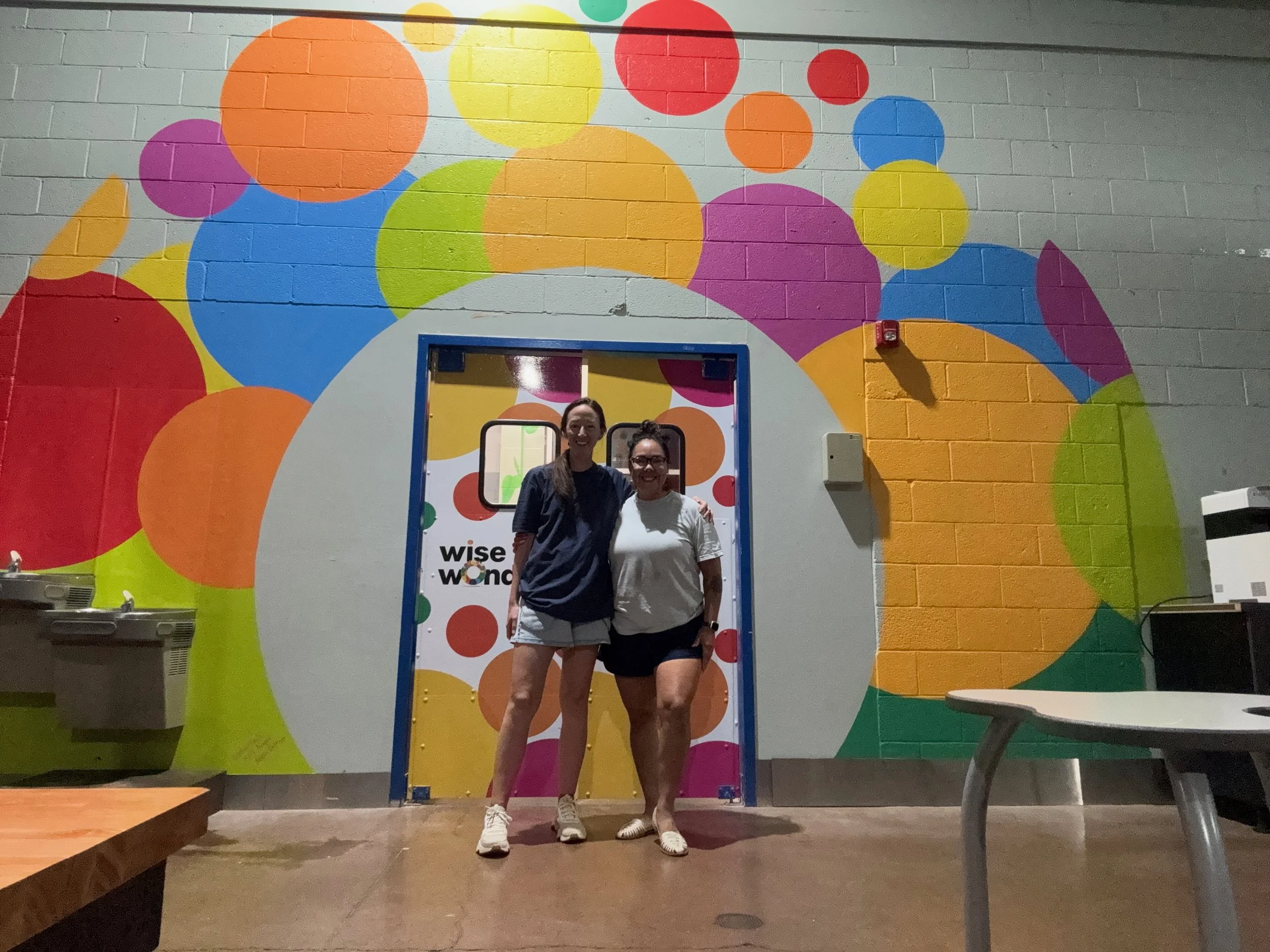 Two women standing inside a colorful mural backdrop at a 'Wise + Wonder' event, smiling and posing for the photo.