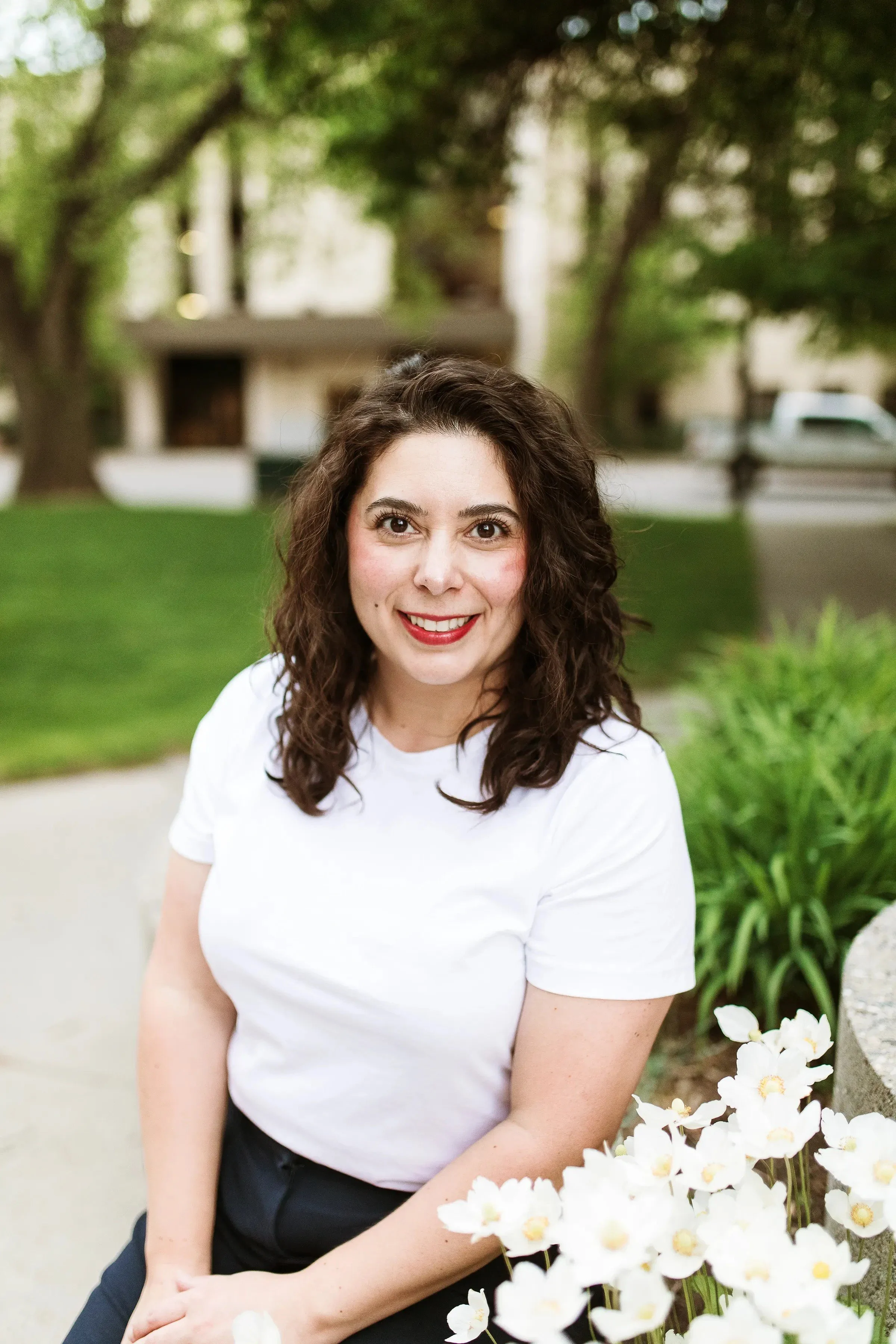 A woman with dark, curly hair smiling at the camera while sitting outdoors near white flowers, trees, and a building in the background.