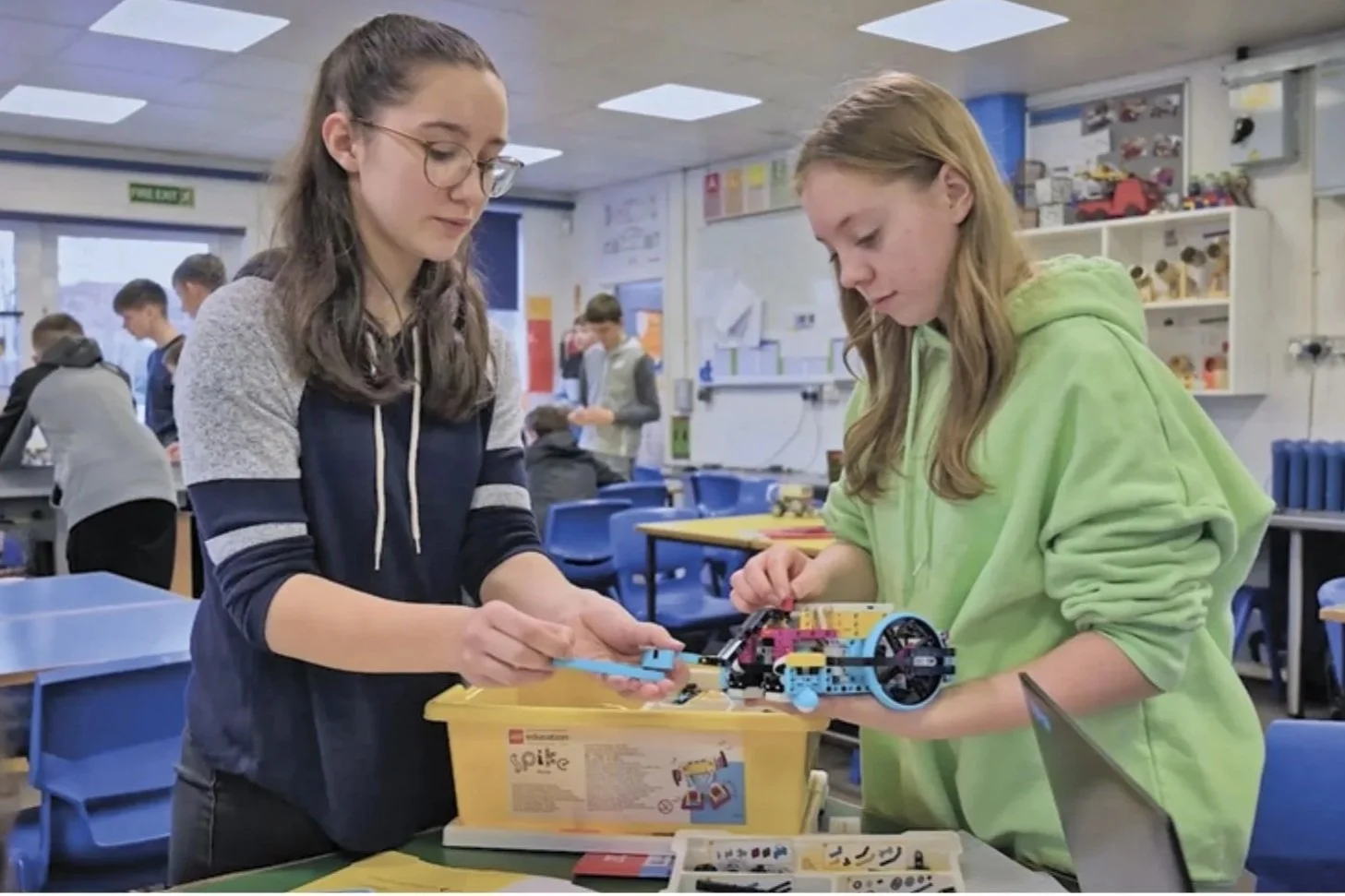 Two girls assembling a Lego robotics kit at a school classroom table, with other students and classroom decorations visible in the background.